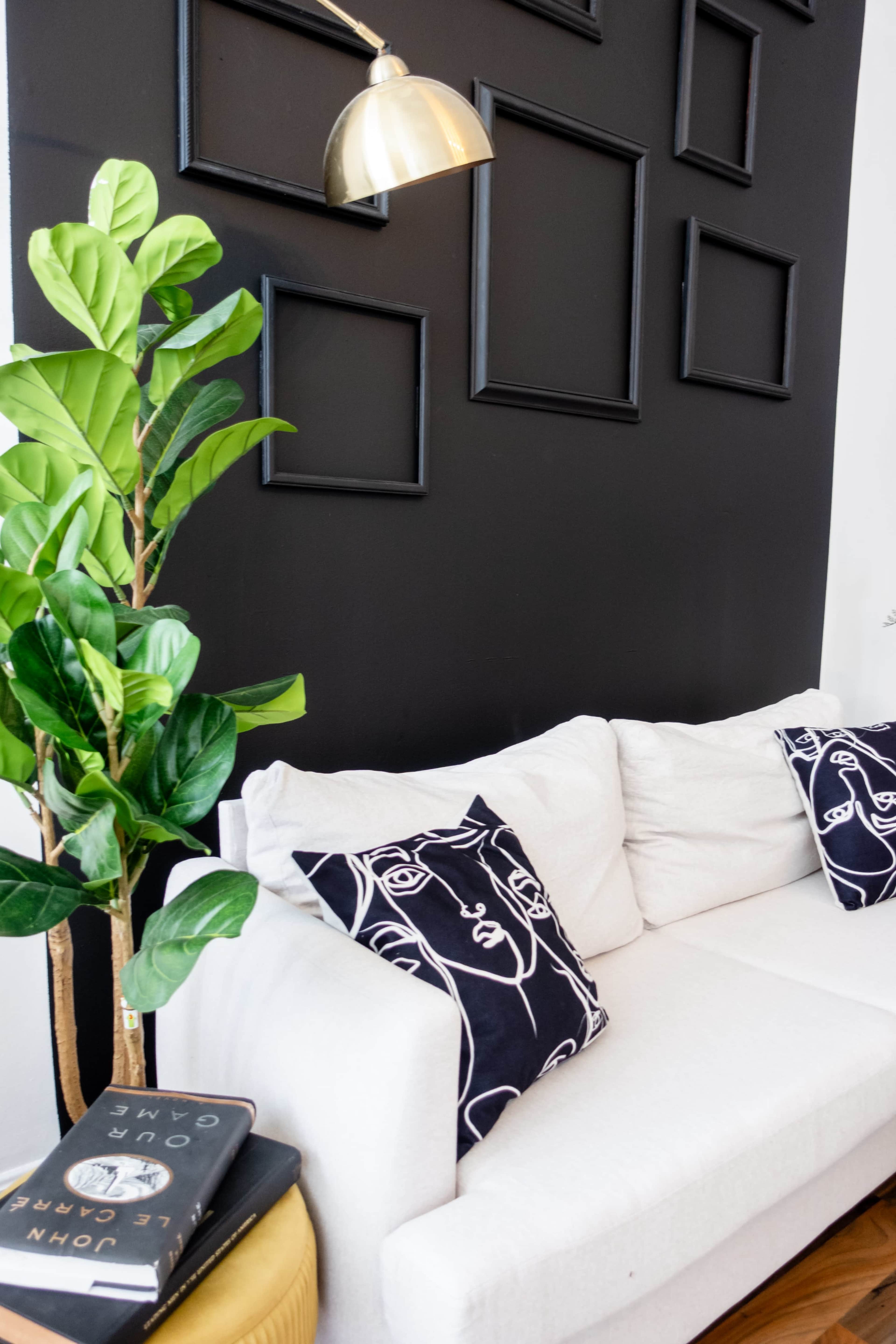 A white sofa is positioned against a black wall adorned with empty frames, while a potted plant stands nearby and books lie on a small table.