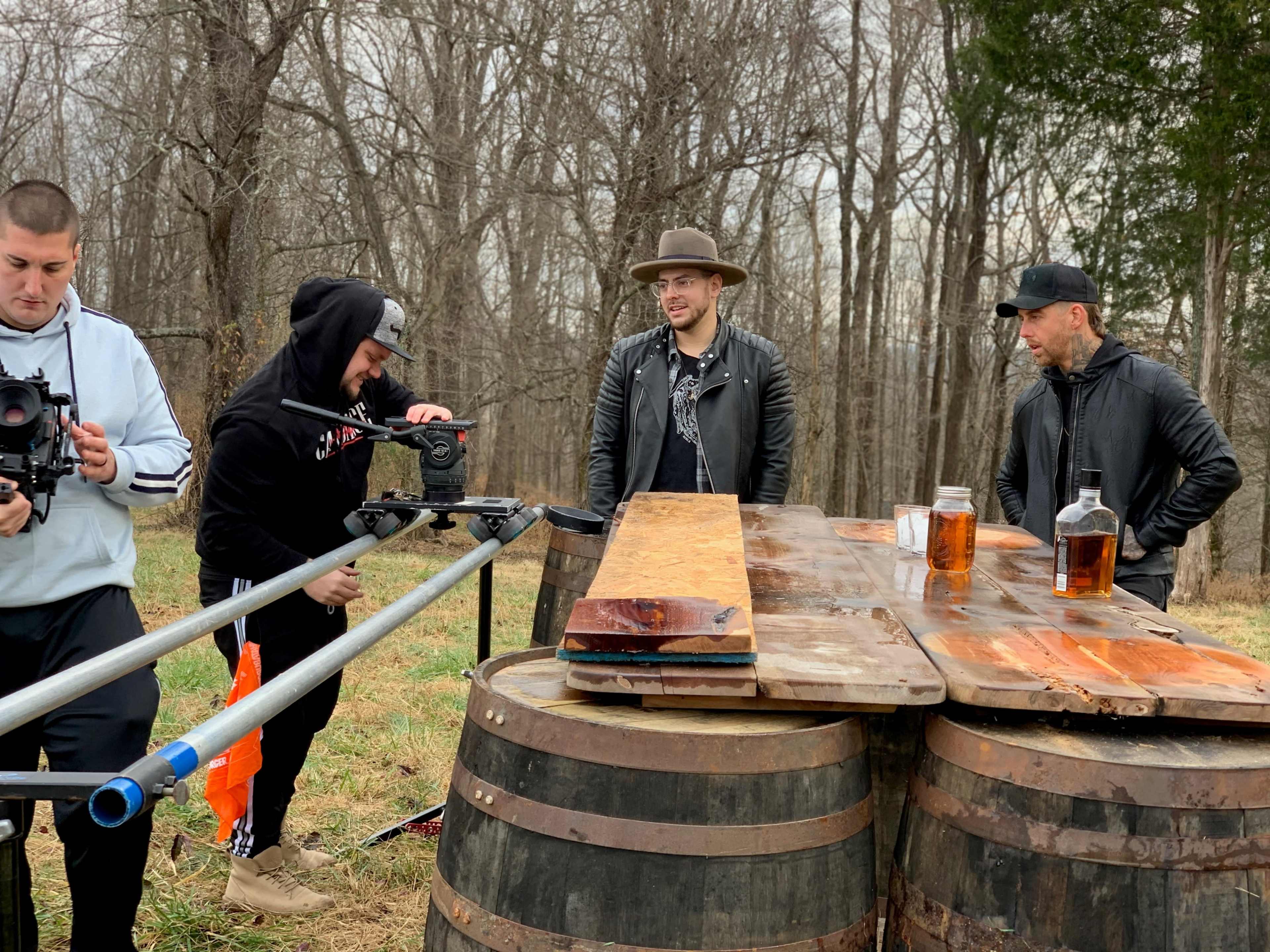 A group of four individuals sets up filming equipment by a rustic wooden table placed on top of two barrels in a wooded area.