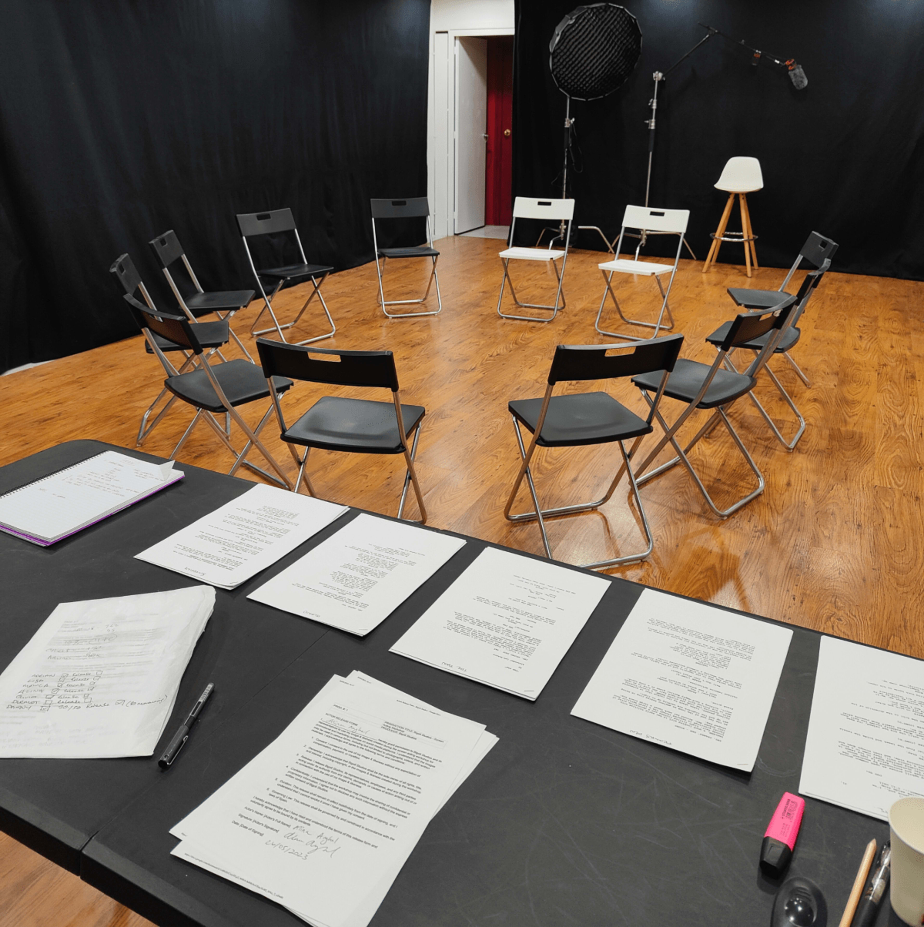 The image shows a rehearsal space with a circular arrangement of black folding chairs on a wooden floor, accompanied by a table covered with scripts and paperwork.