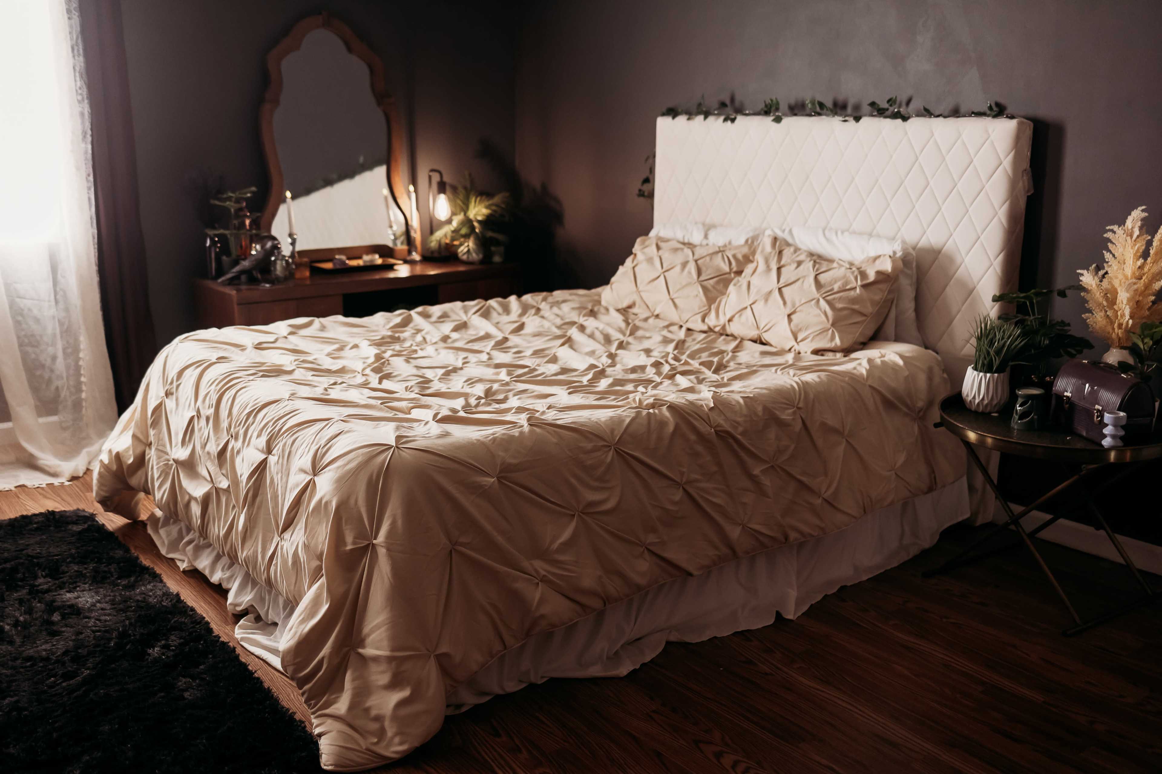 A neatly made bed with a textured beige comforter is positioned against a dark wall, accompanied by a mirrored dresser and a plant on a side table.