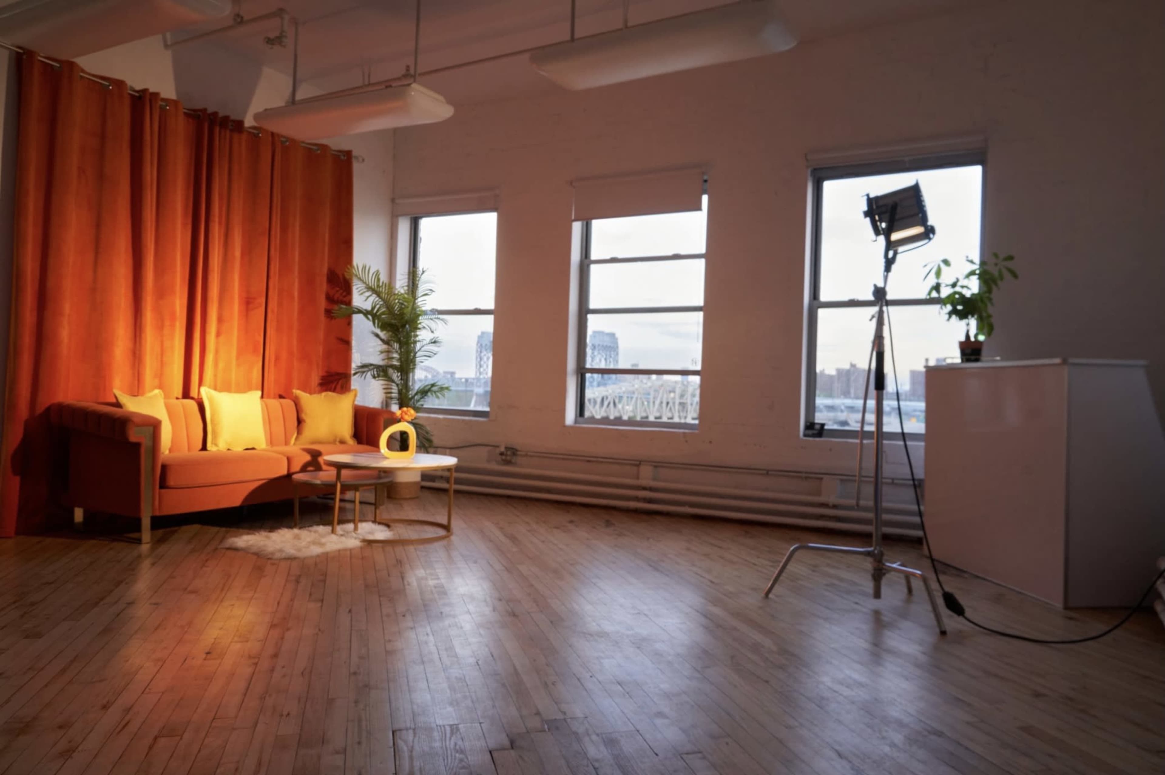 A cozy living space features an orange couch with yellow cushions, a small round rug, and a lamp, illuminated by natural light from large windows.