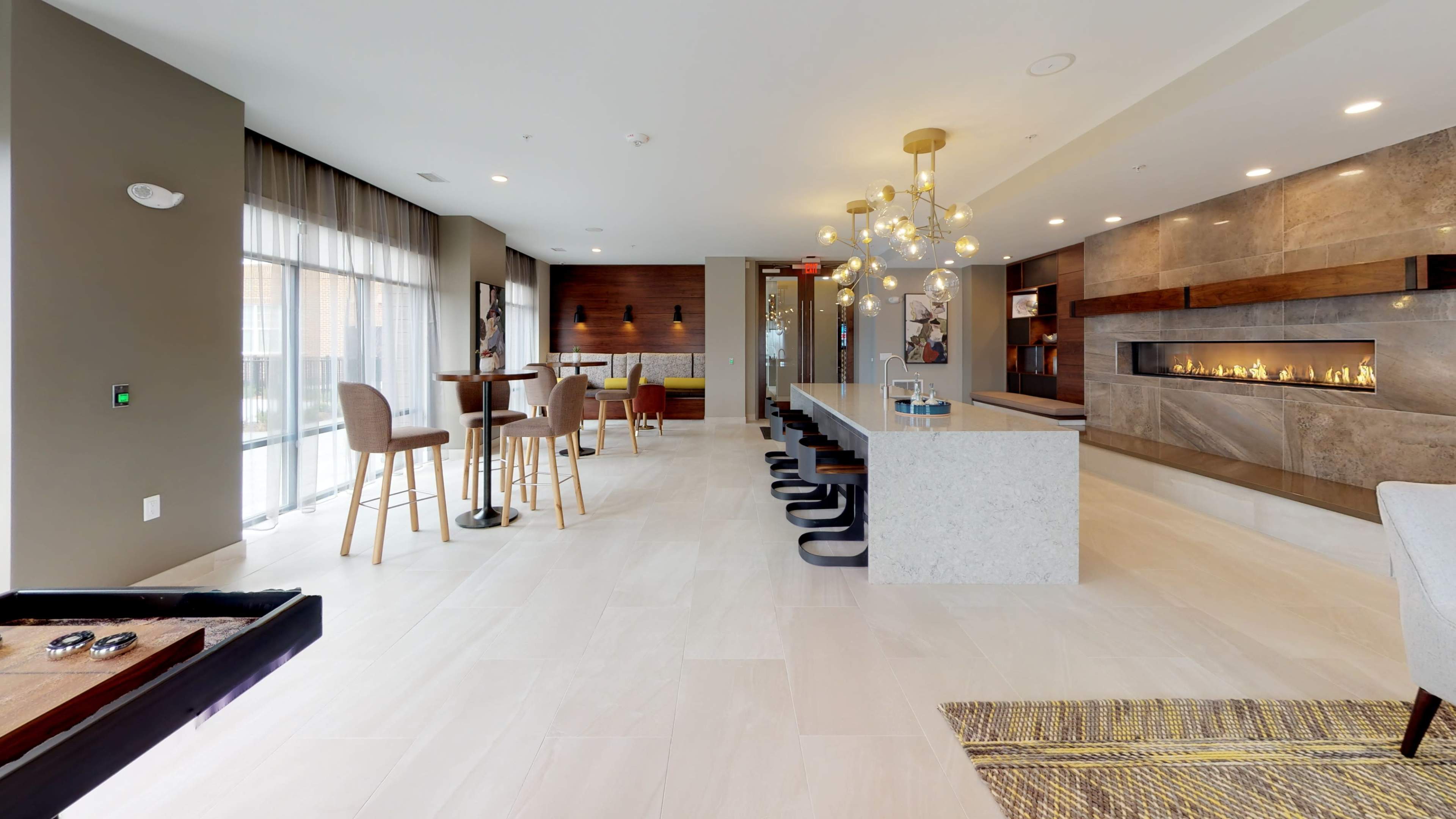 The image shows a modern communal lounge area featuring a long countertop, barstools, a small dining table, and a fireplace along the wall.