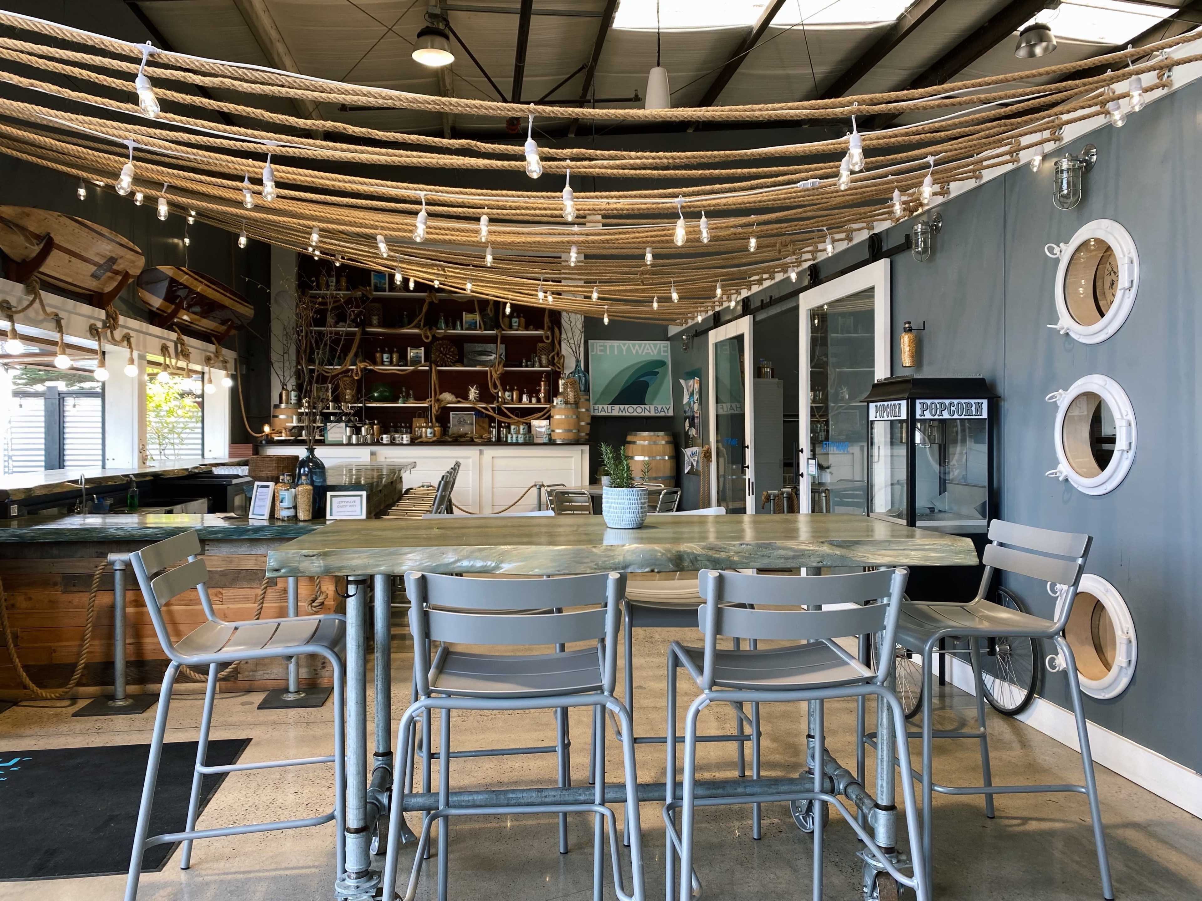A nautical-themed bar area features a metal table surrounded by four gray stools, with decorative ropes and lights overhead.