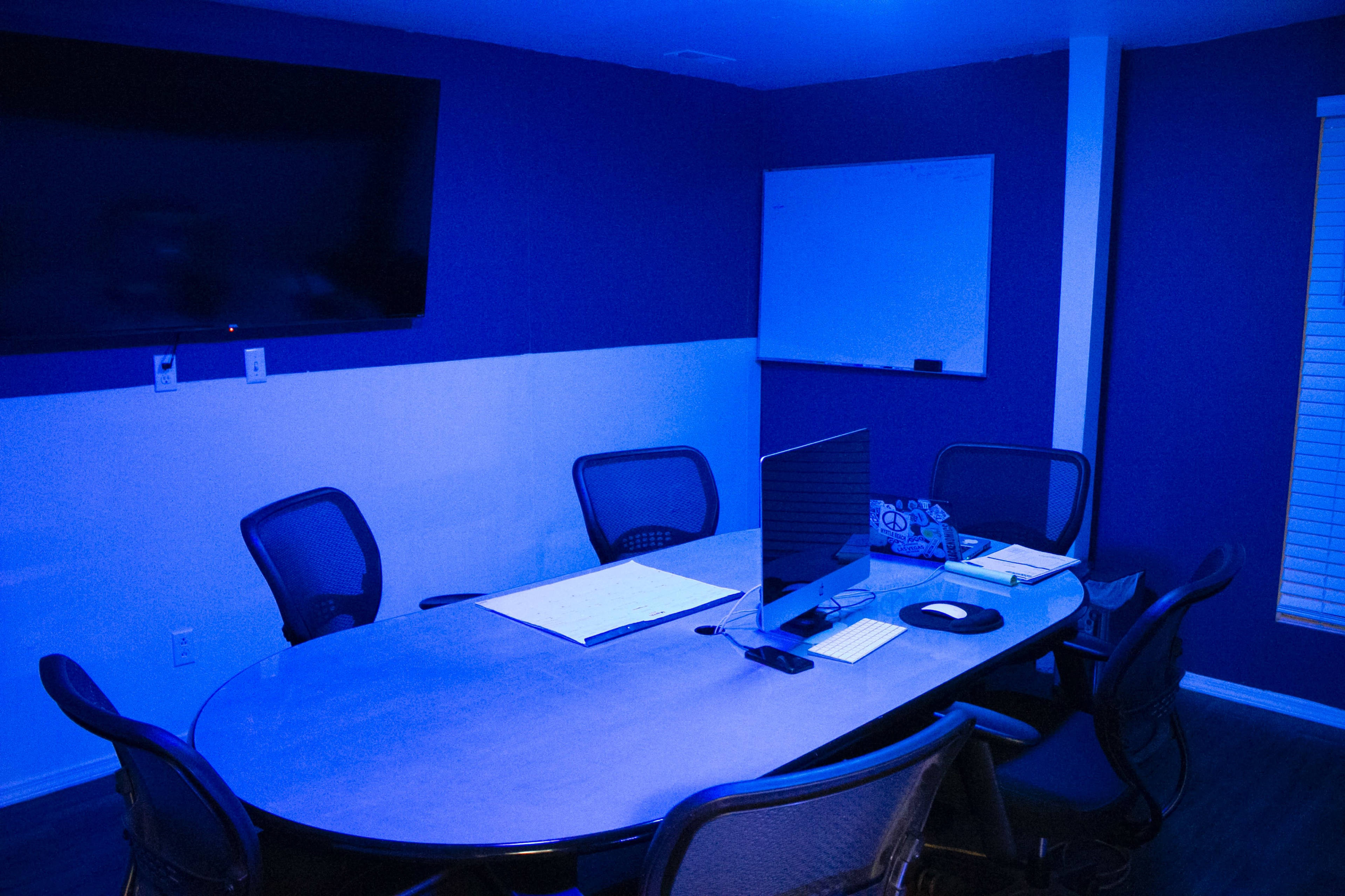 A dimly lit conference room features a large oval table with several chairs, a computer setup, a whiteboard on the wall, and a large screen mounted above.
