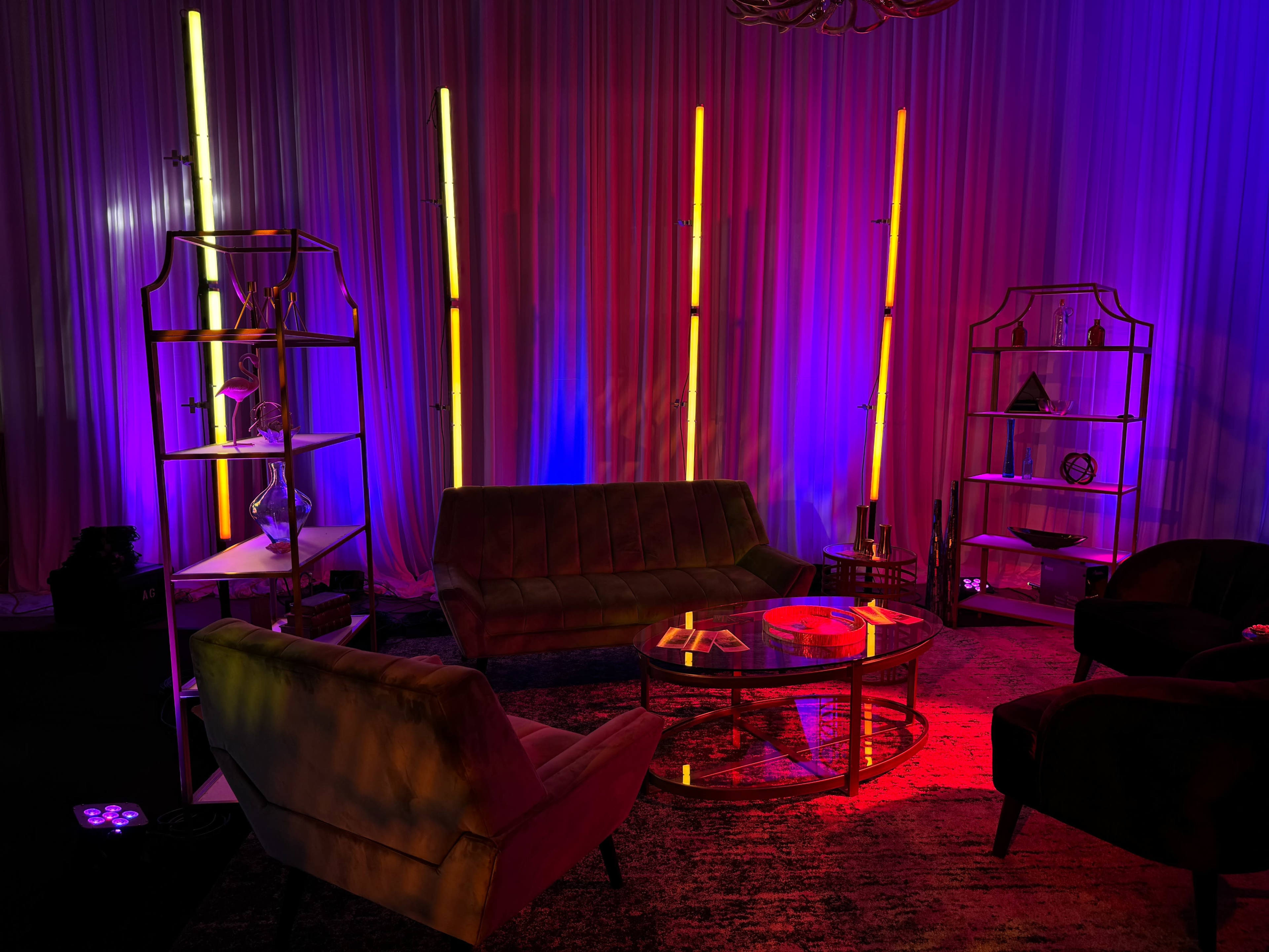 A modern lounge area features a green sofa, armchairs, a glass coffee table, and illuminated shelves against a backdrop of colorful lighting and draped fabric.