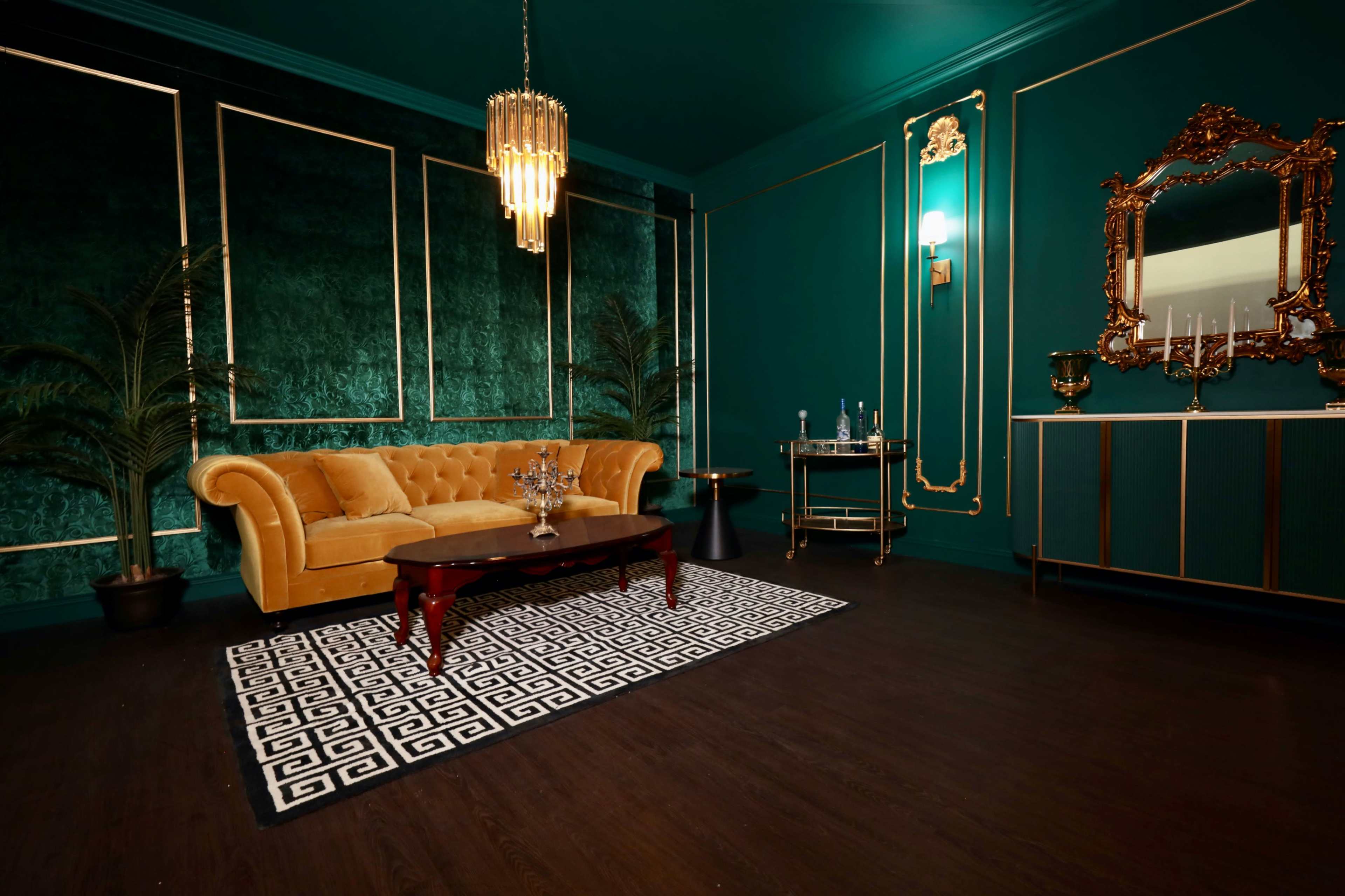 A stylish room featuring a green velvet wall, a yellow tufted sofa, a coffee table, and a decorative mirror, illuminated by a chandelier.
