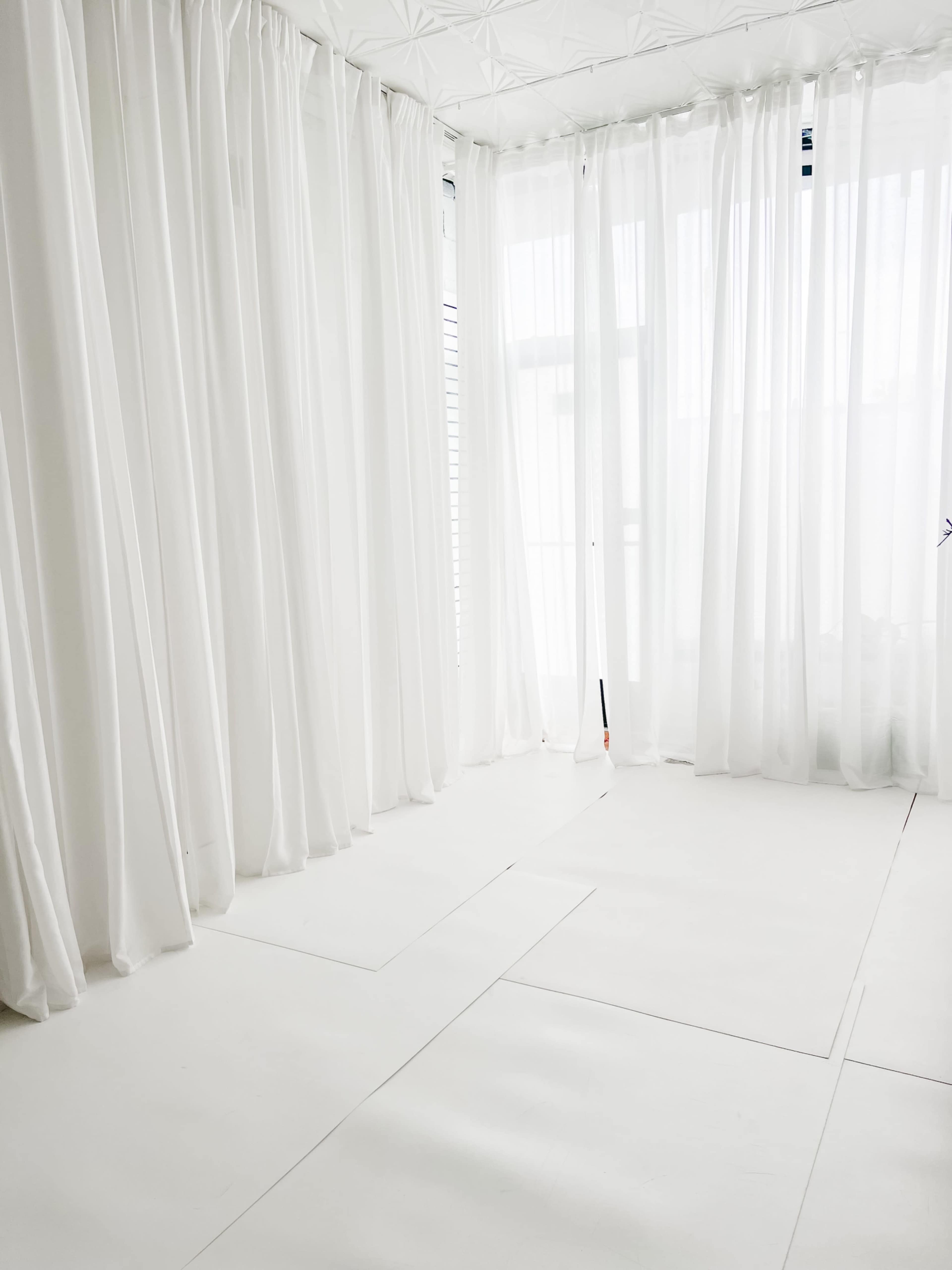 A bright, empty room features white walls and floor, with sheer white curtains allowing natural light to enter.