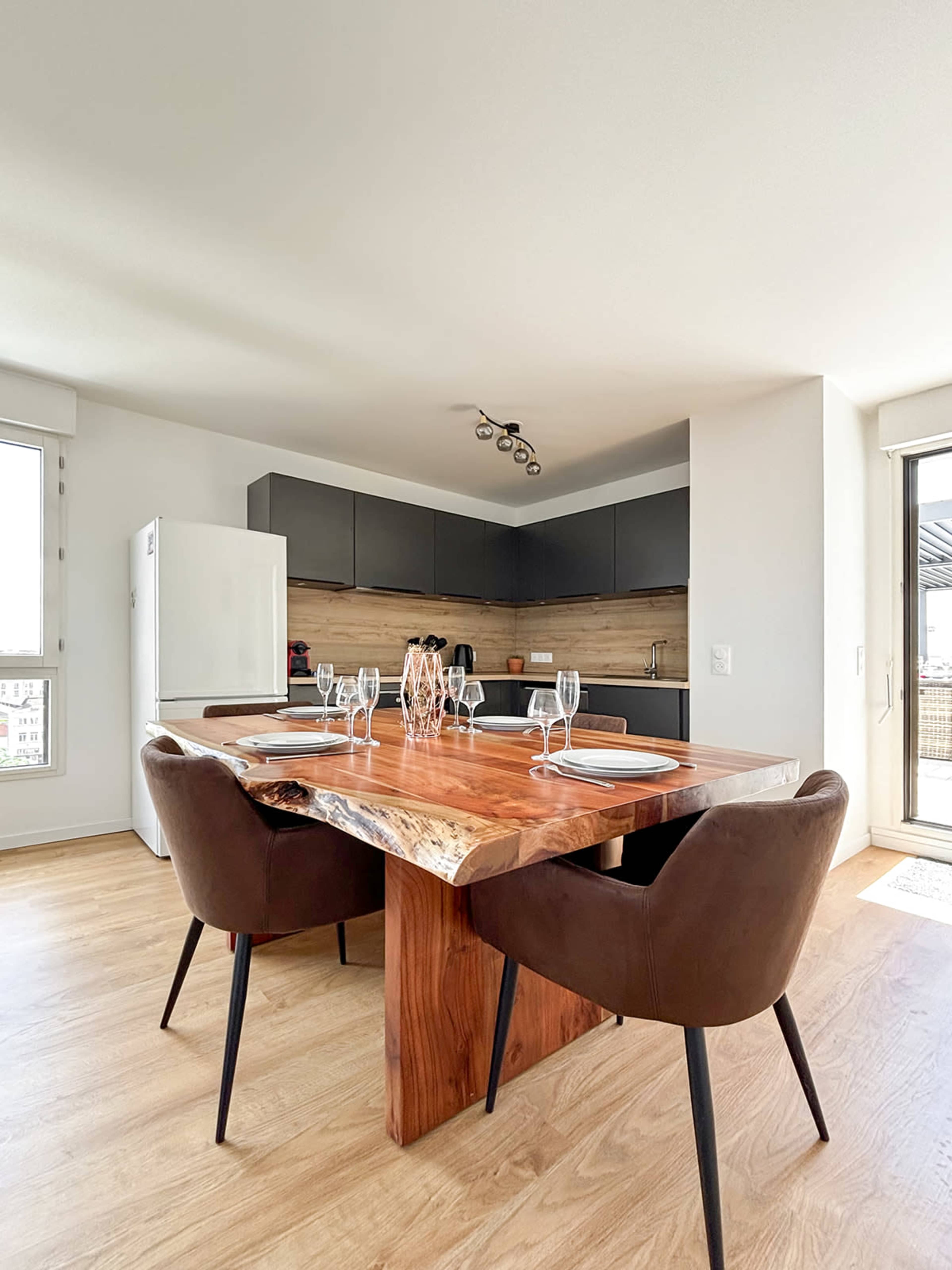 A modern kitchen features a wooden dining table with four chairs, set with plates and glasses, alongside sleek cabinetry and a large window.