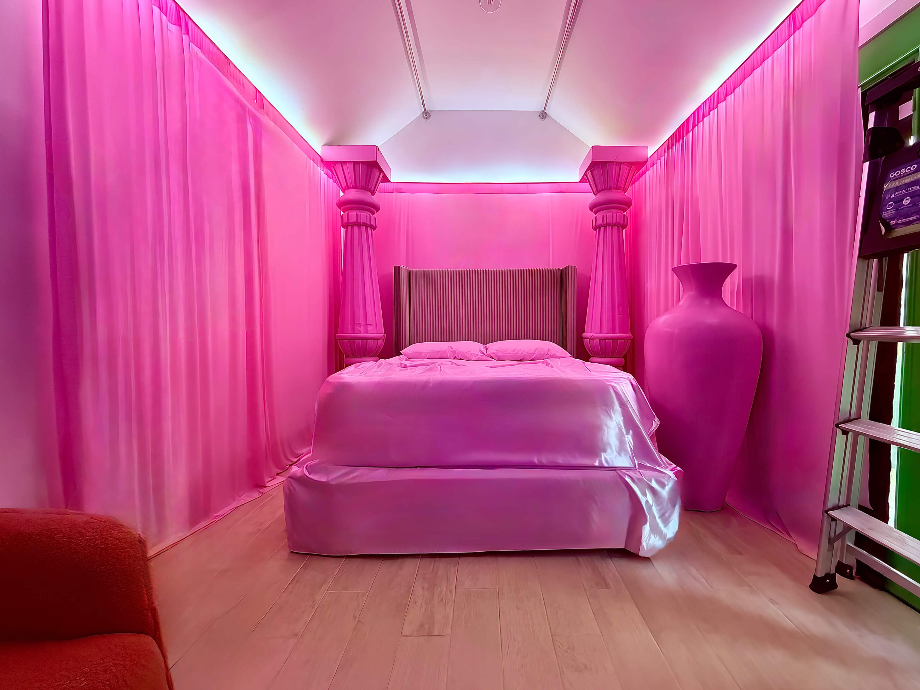 The image shows a bedroom draped in pink fabric, featuring a large bed, a tall pink vase, and soft lighting emanating from the walls.