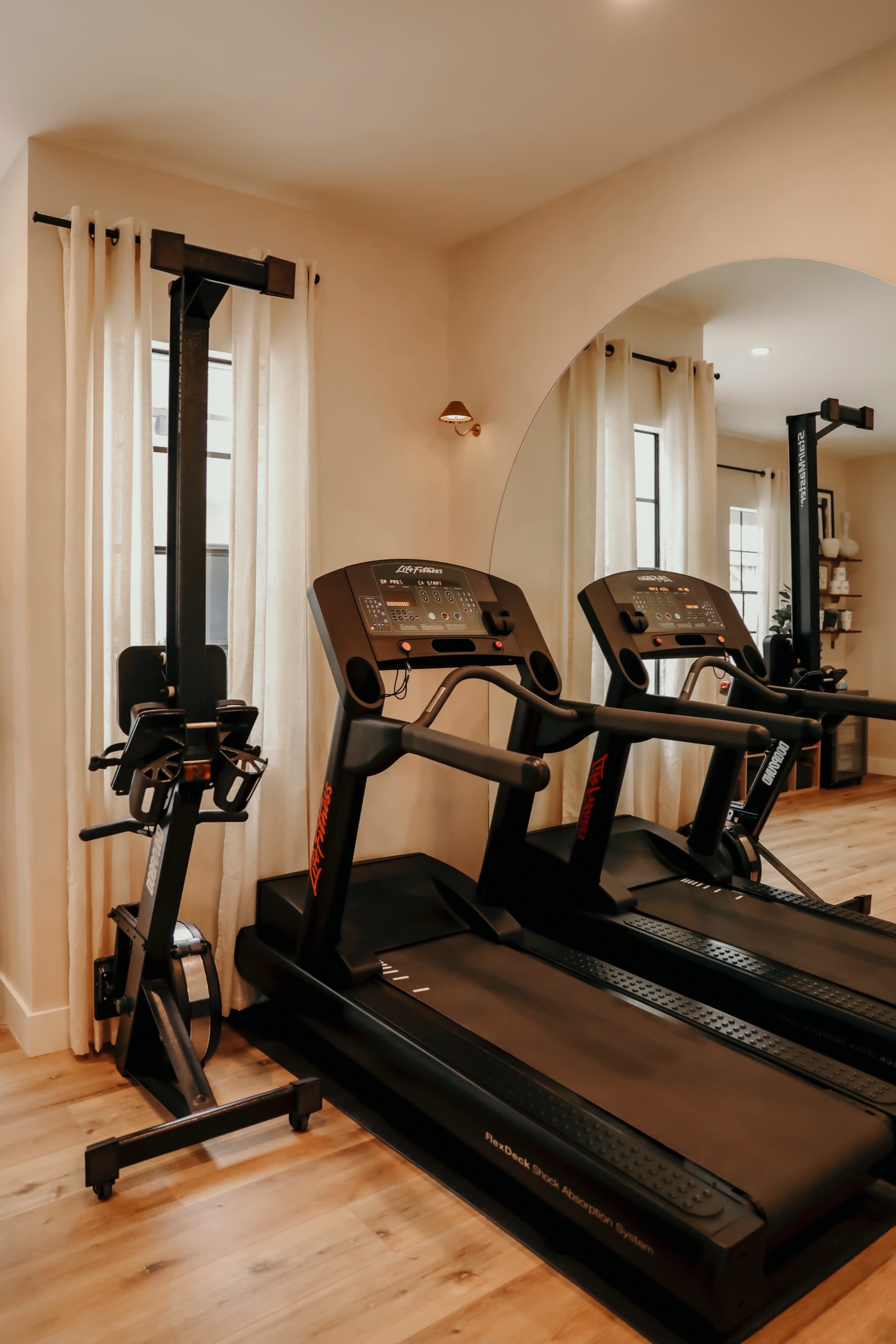 The image shows a well-lit home gym featuring two treadmills and a strength training machine against a wall with light-colored curtains.