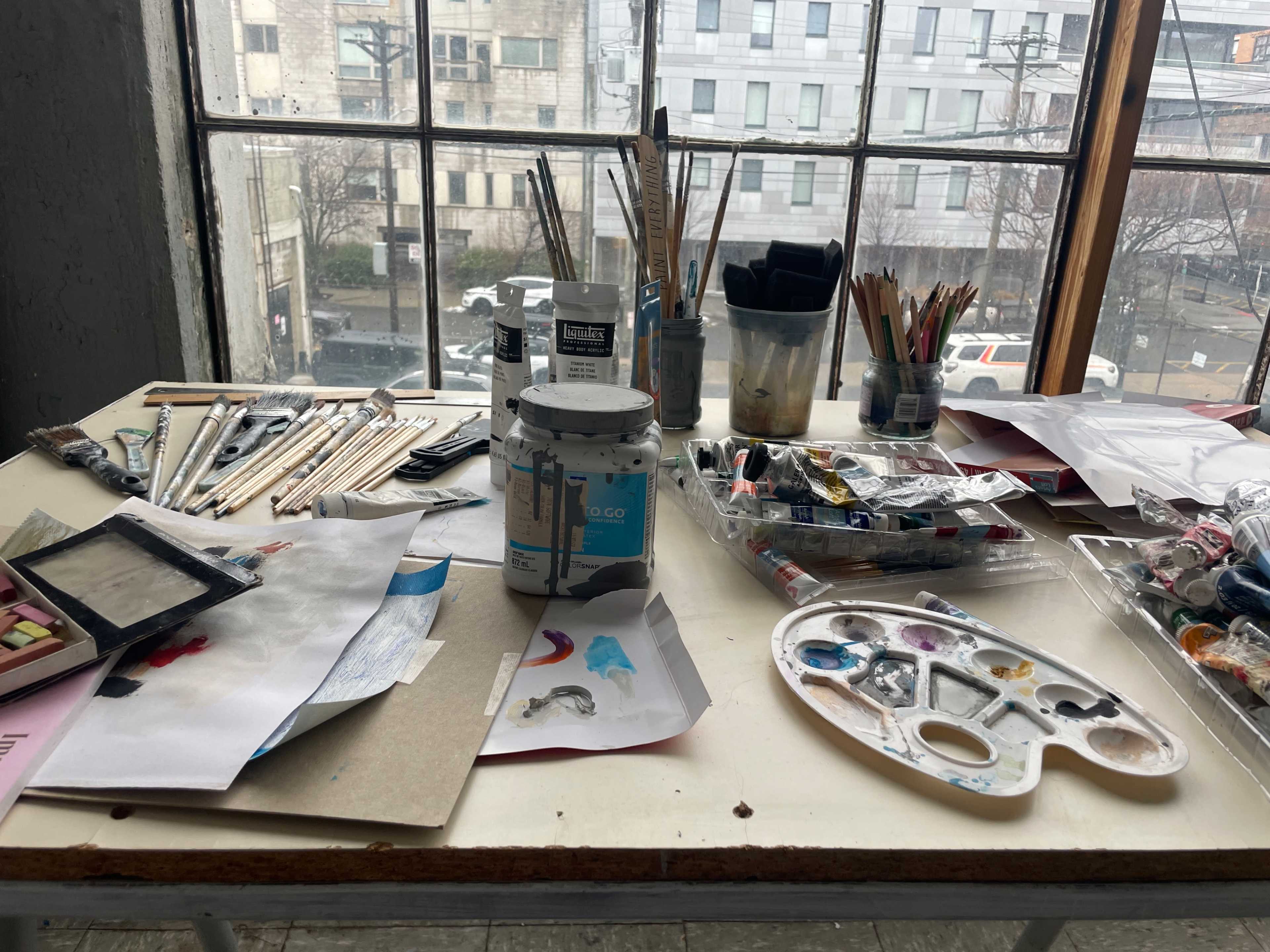 The image shows a cluttered artist's workspace featuring various art supplies, paint tubes, a palette, and papers scattered across a table near a large window.