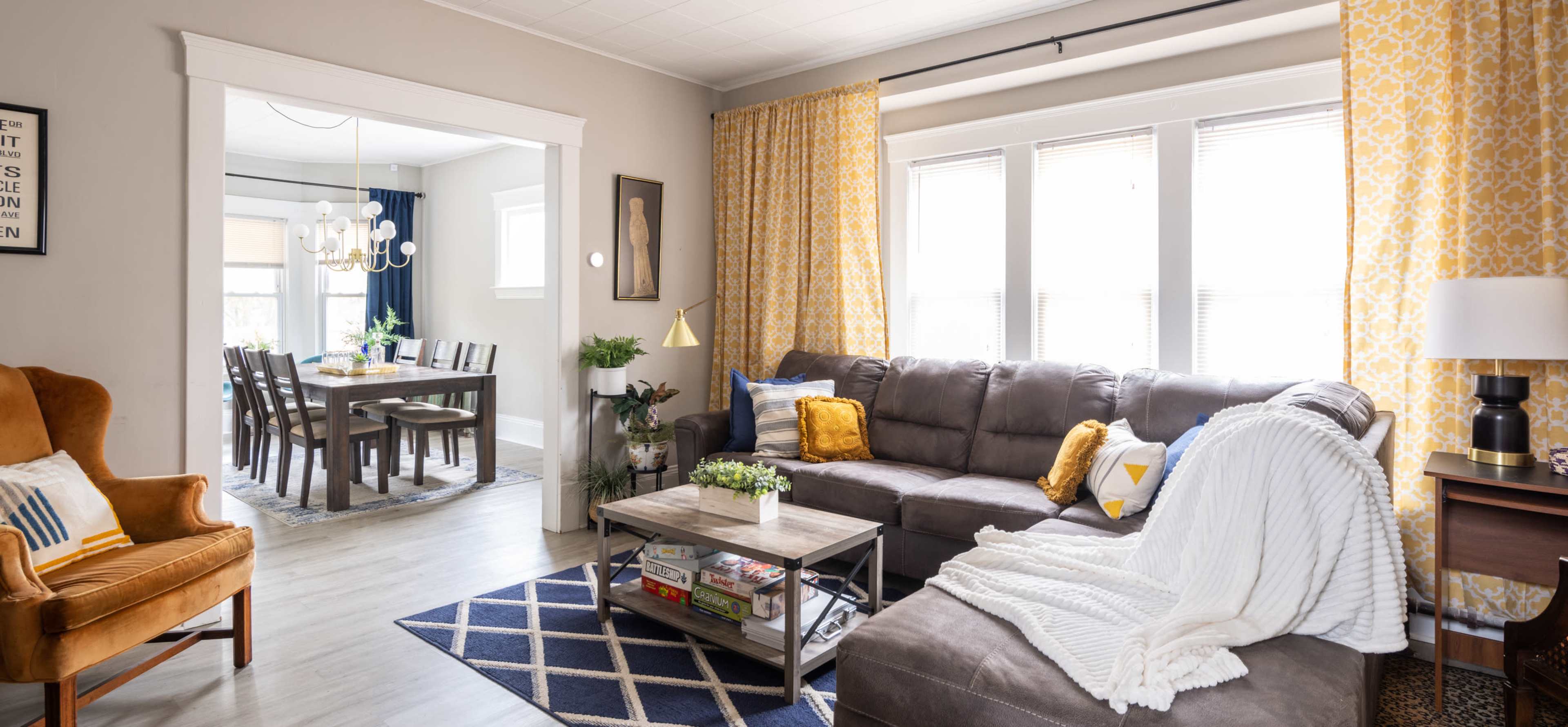 A living room features a grey sofa, a coffee table with books, and yellow curtains that open to a dining area with a large table and chairs.