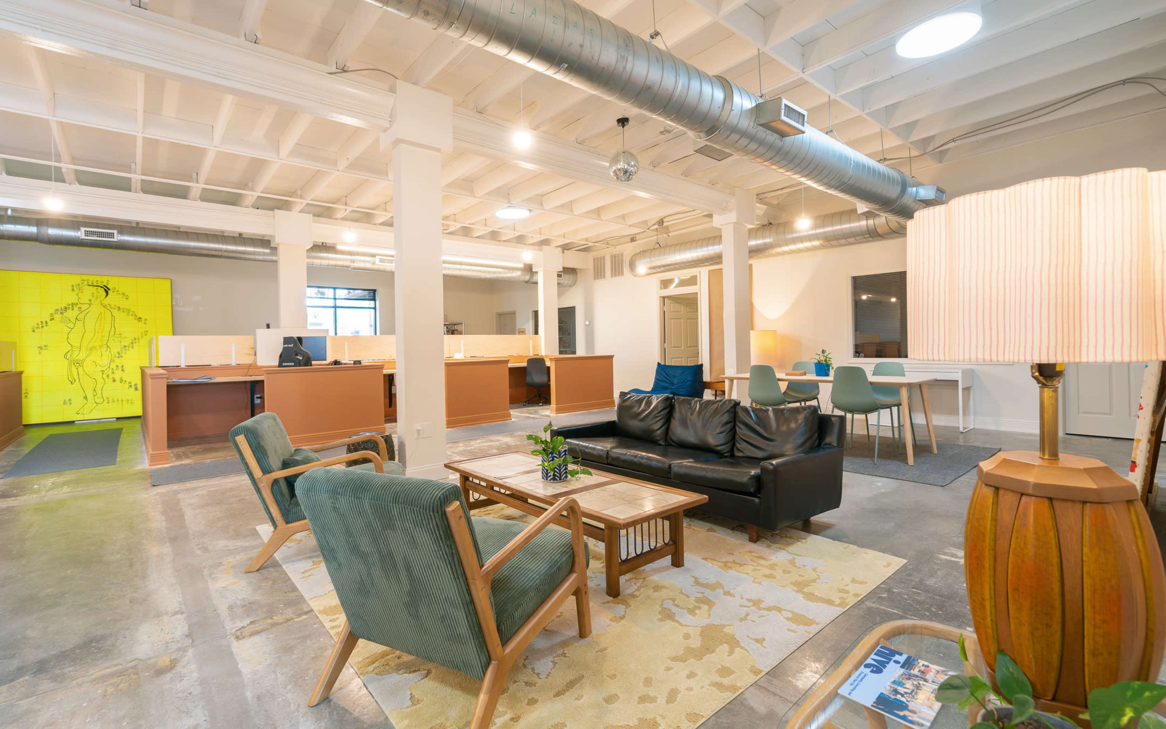 A modern office space features a seating area with a black sofa, wooden chairs, and a coffee table, alongside workstations in a bright, open layout.
