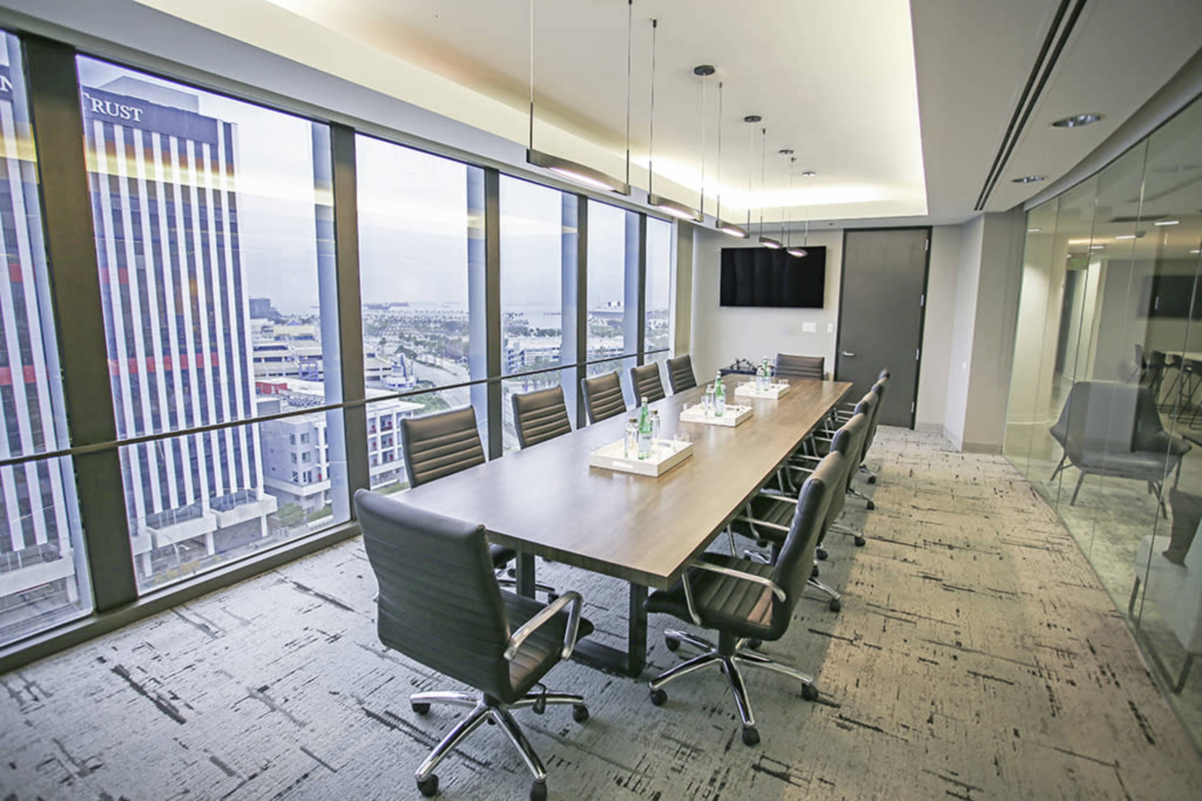 A spacious conference room features a long wooden table surrounded by ergonomic chairs, with large windows offering a view of the city skyline.