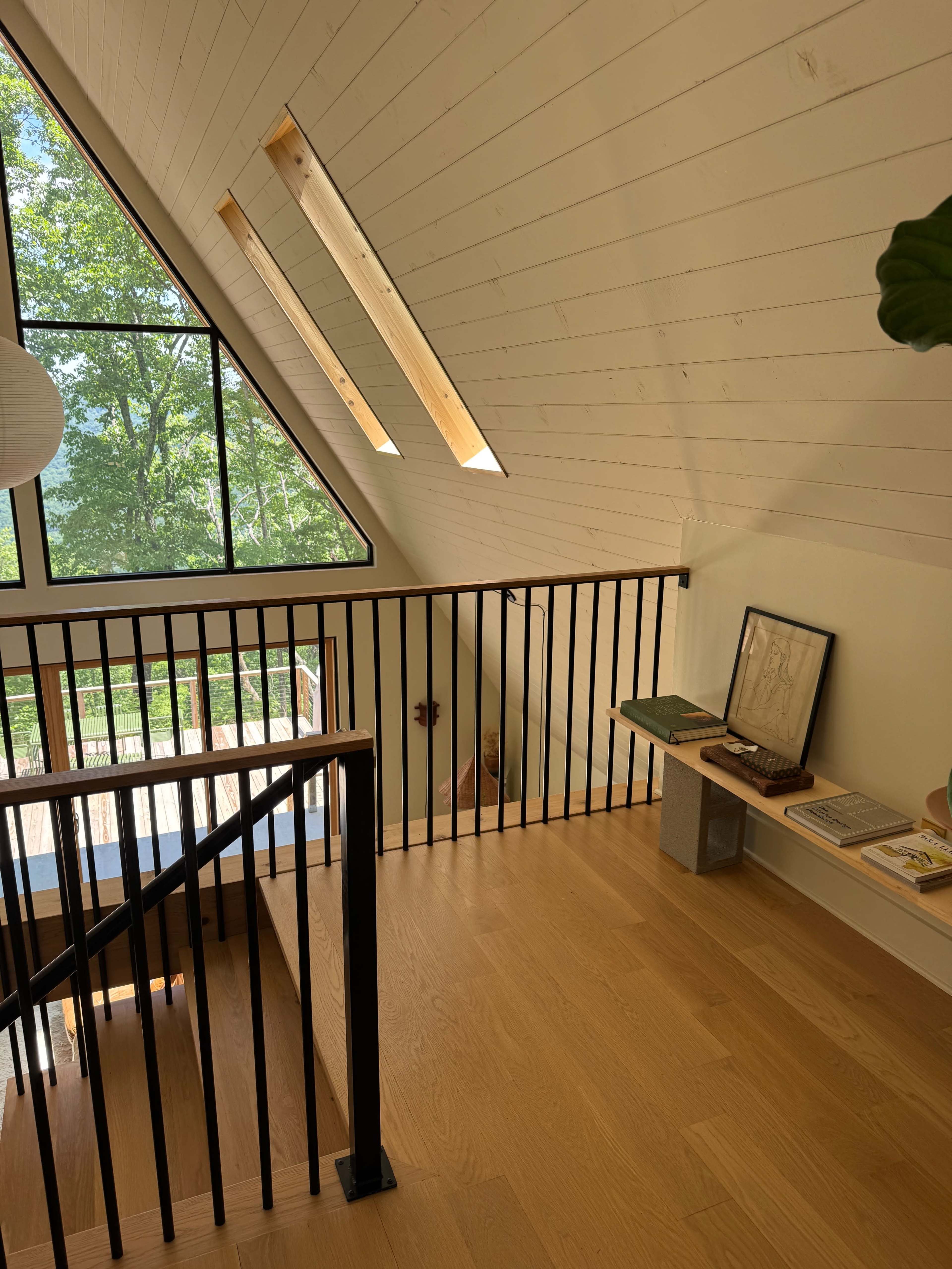 The image shows the interior of a lofted space with an angled ceiling, wooden flooring, and a railing alongside a staircase, leading to a window with views of greenery outside.