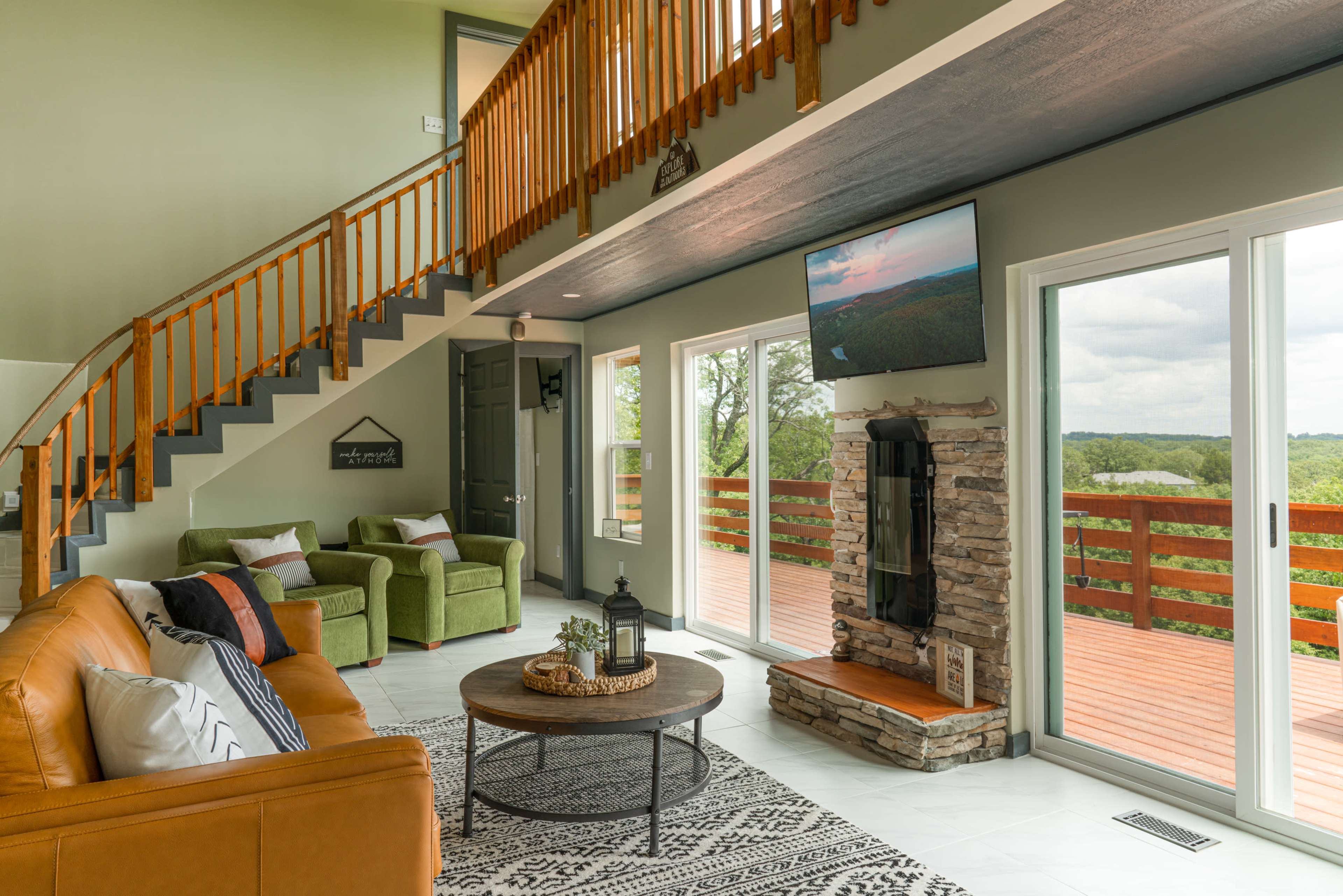 A spacious living room with a leather sofa and green armchairs, a stone fireplace, a round coffee table, and large windows overlooking a deck and landscape beyond.