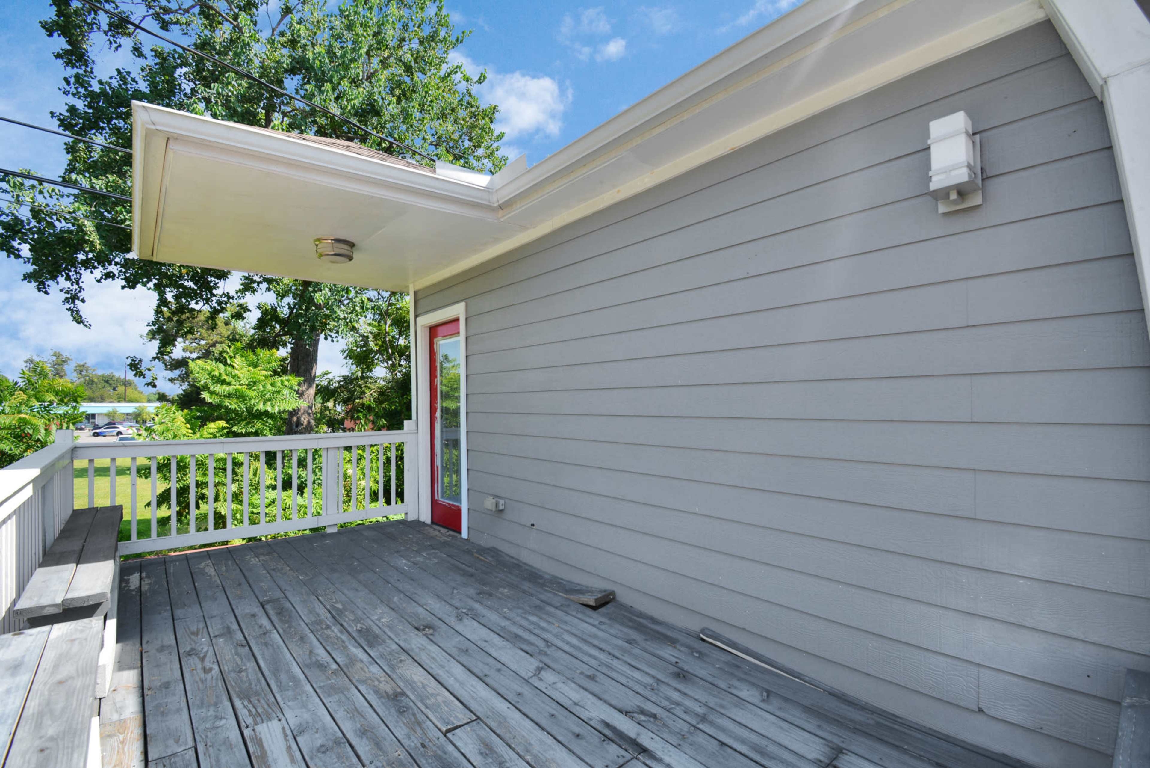 A wooden deck beside a light gray house with a red door and lush green trees in the background.