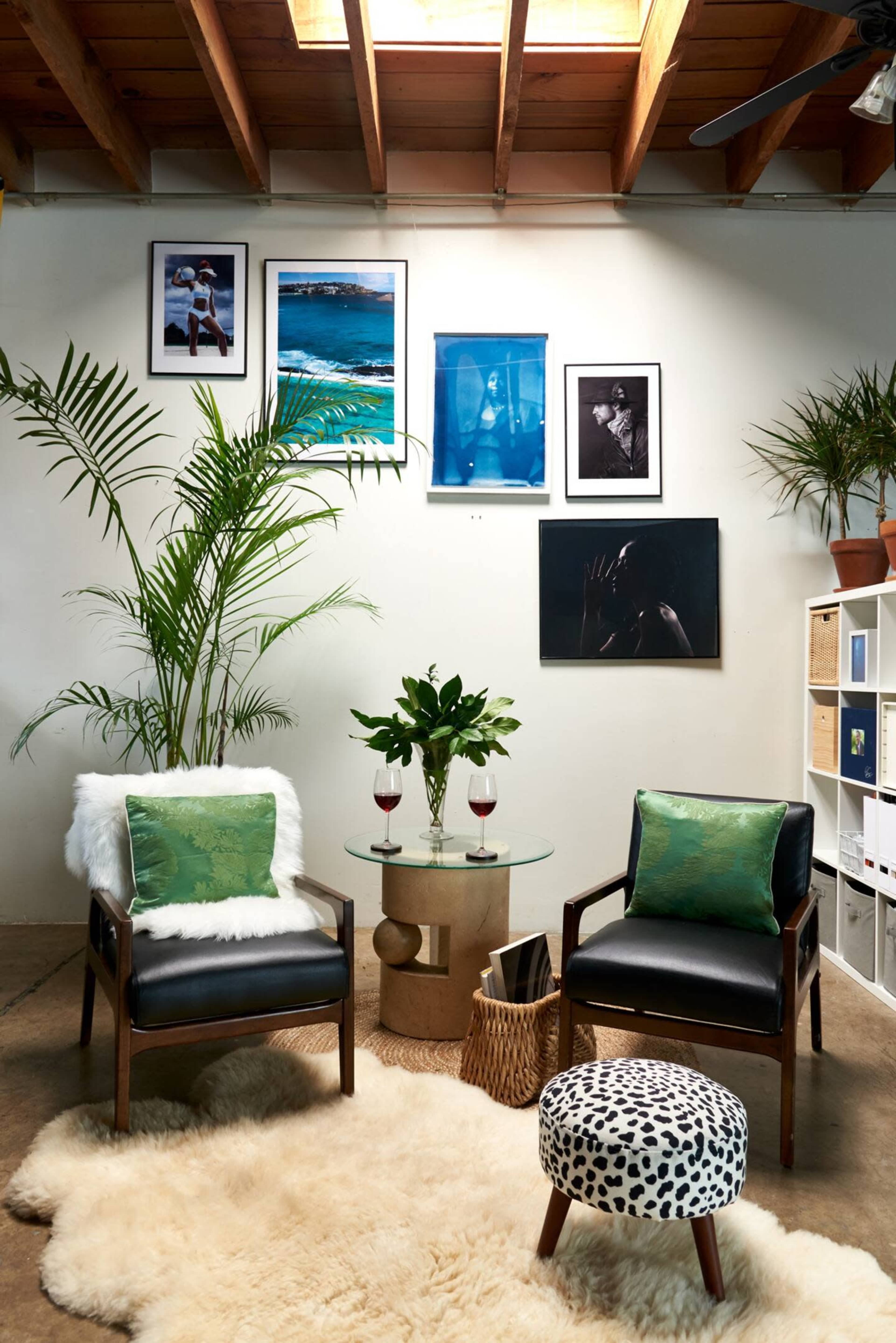 The scene features two black lounge chairs with green cushions, a circular glass table with two glasses of red drinks, a patterned ottoman, and various framed artworks on the wall, all set against a backdrop of potted plants and a textured rug.
