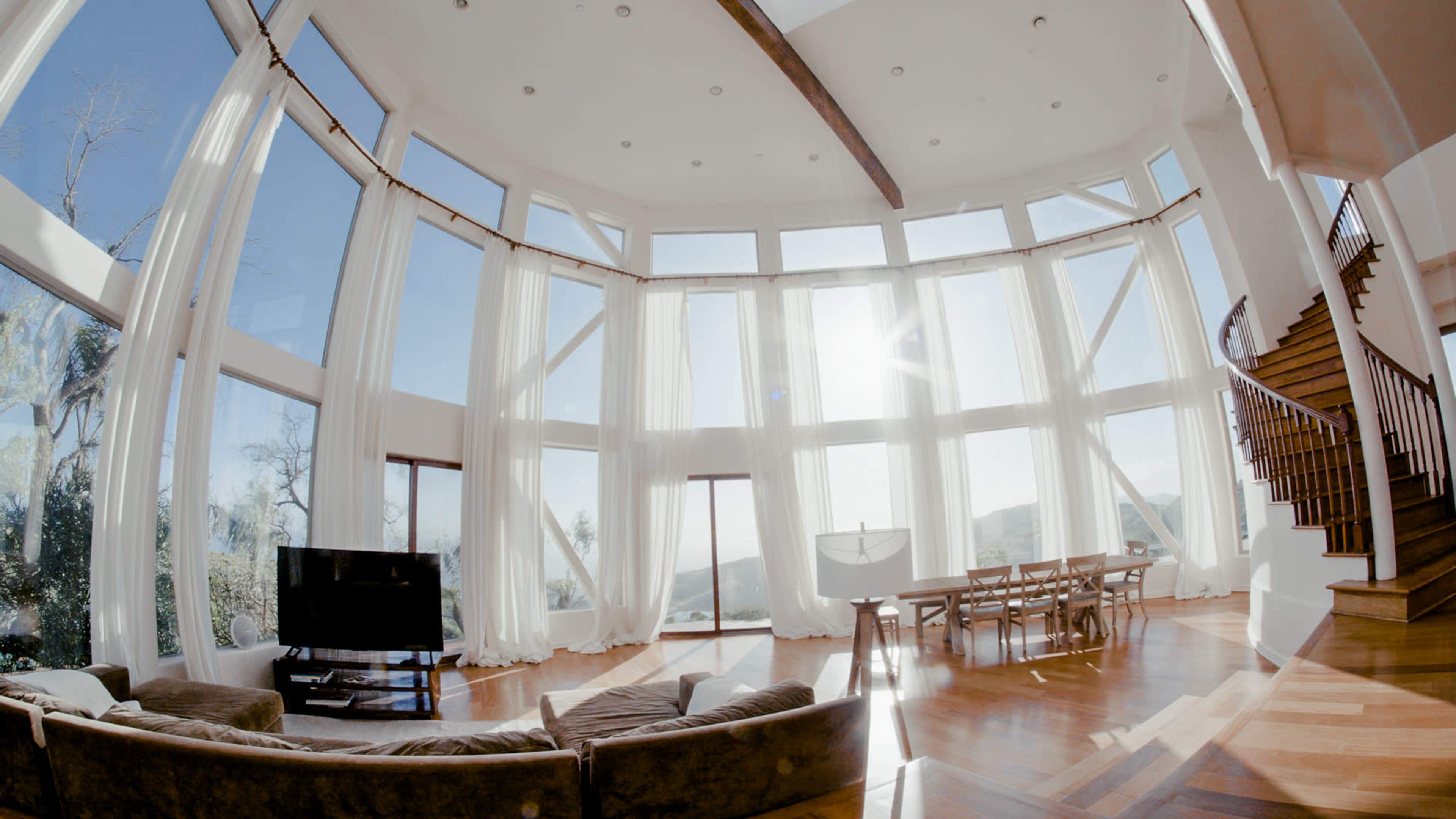 A spacious living room features large windows with sheer curtains, a staircase, a dining area, and a television.
