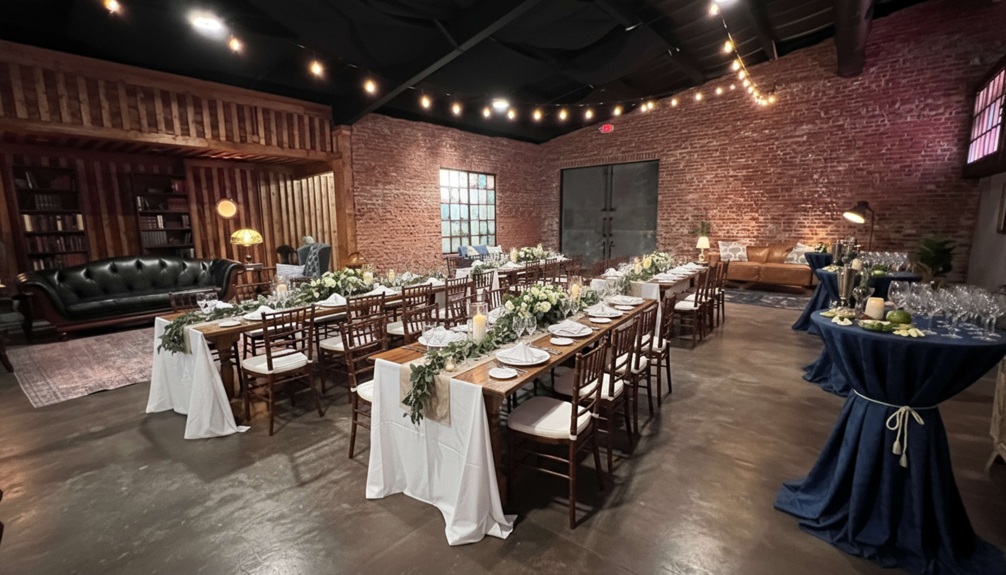 The image shows a banquet setup in a rustic venue featuring wooden tables adorned with white tablecloths, floral centerpieces, and surrounded by wooden chairs under string lights.