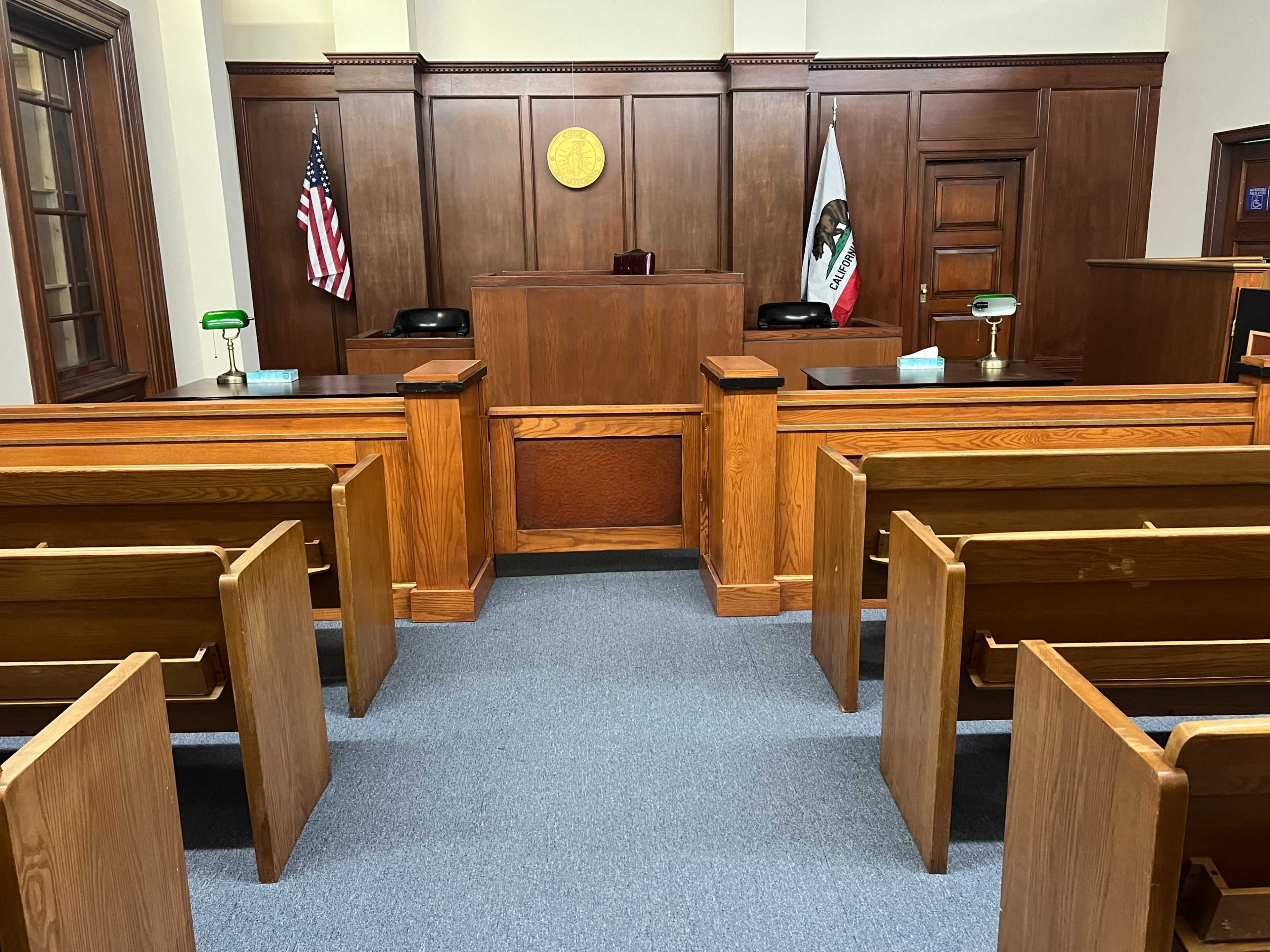 A wooden courtroom with two rows of benches facing a judges' bench and a state flag.
