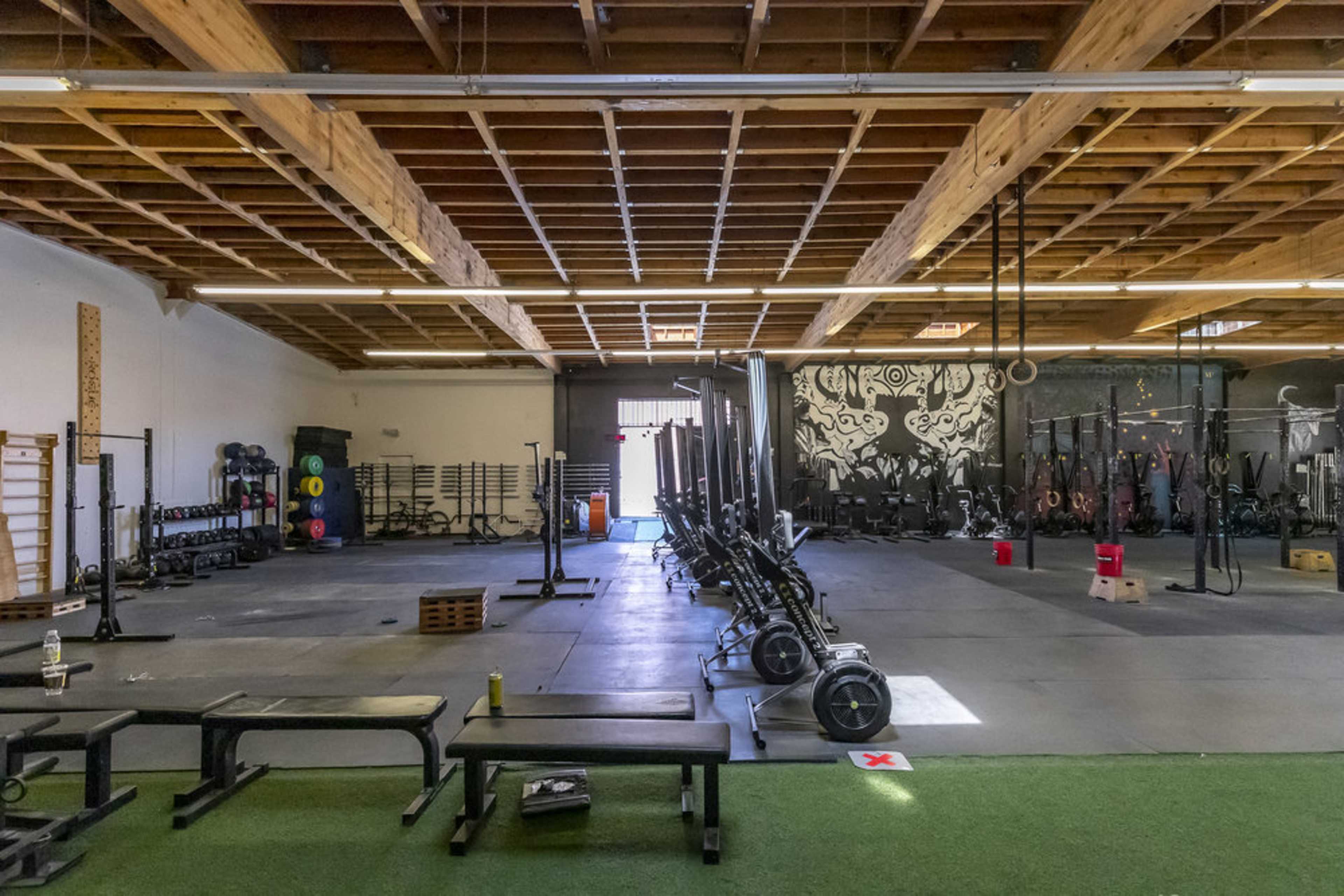 The image displays a spacious gym interior with various workout equipment arranged neatly across the floor, including weightlifting benches, racks, and exercise machines.
