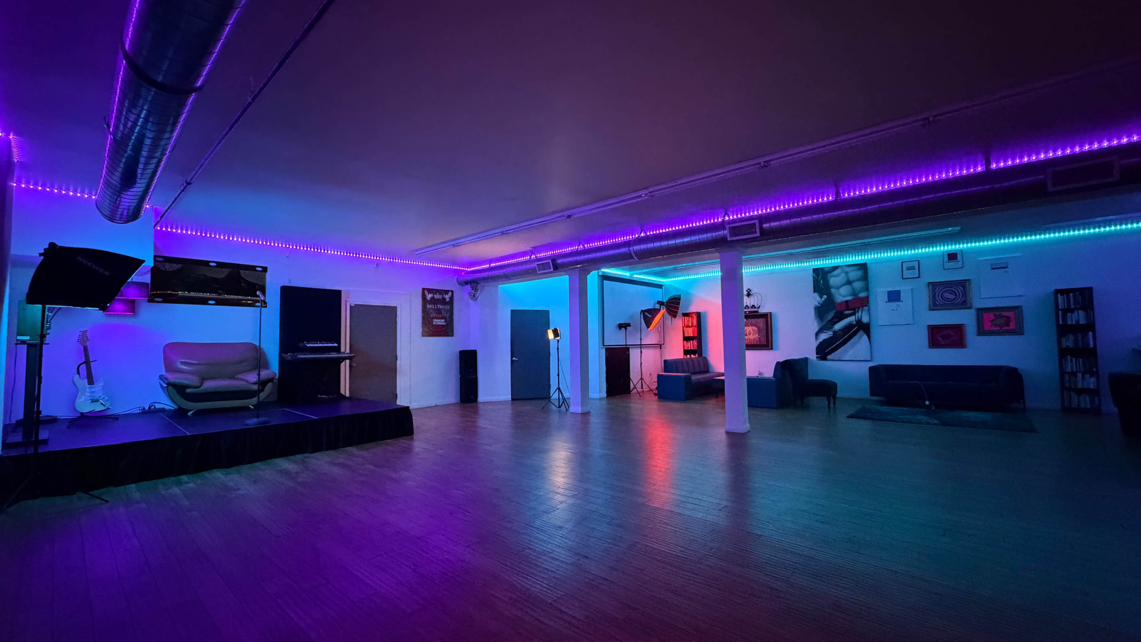 The image shows a spacious room with colorful LED lighting, featuring a small stage, seating arrangements, and an assortment of wall art.