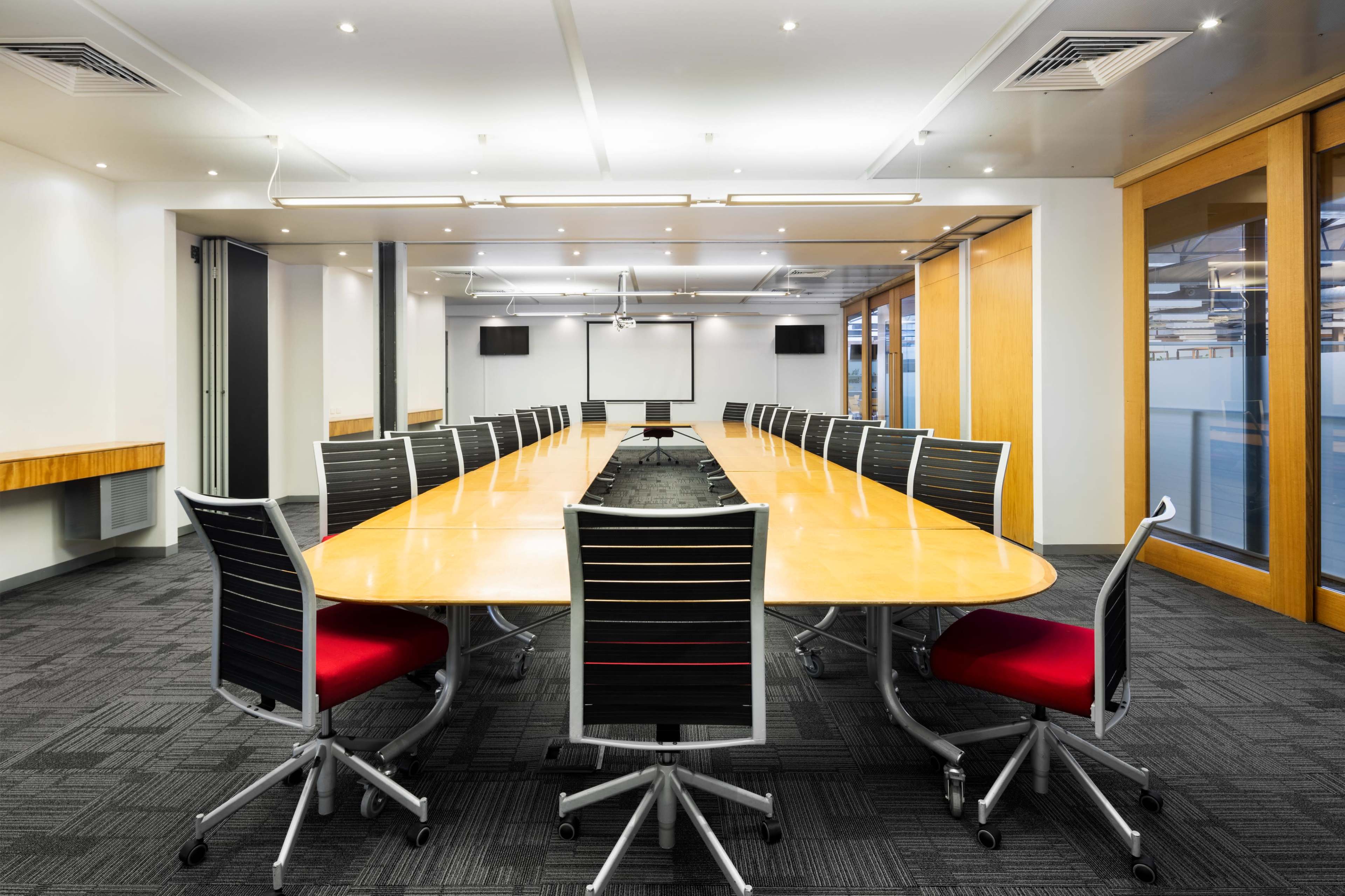A large conference room features a long wooden table surrounded by black chairs with red accents, under bright ceiling lights.