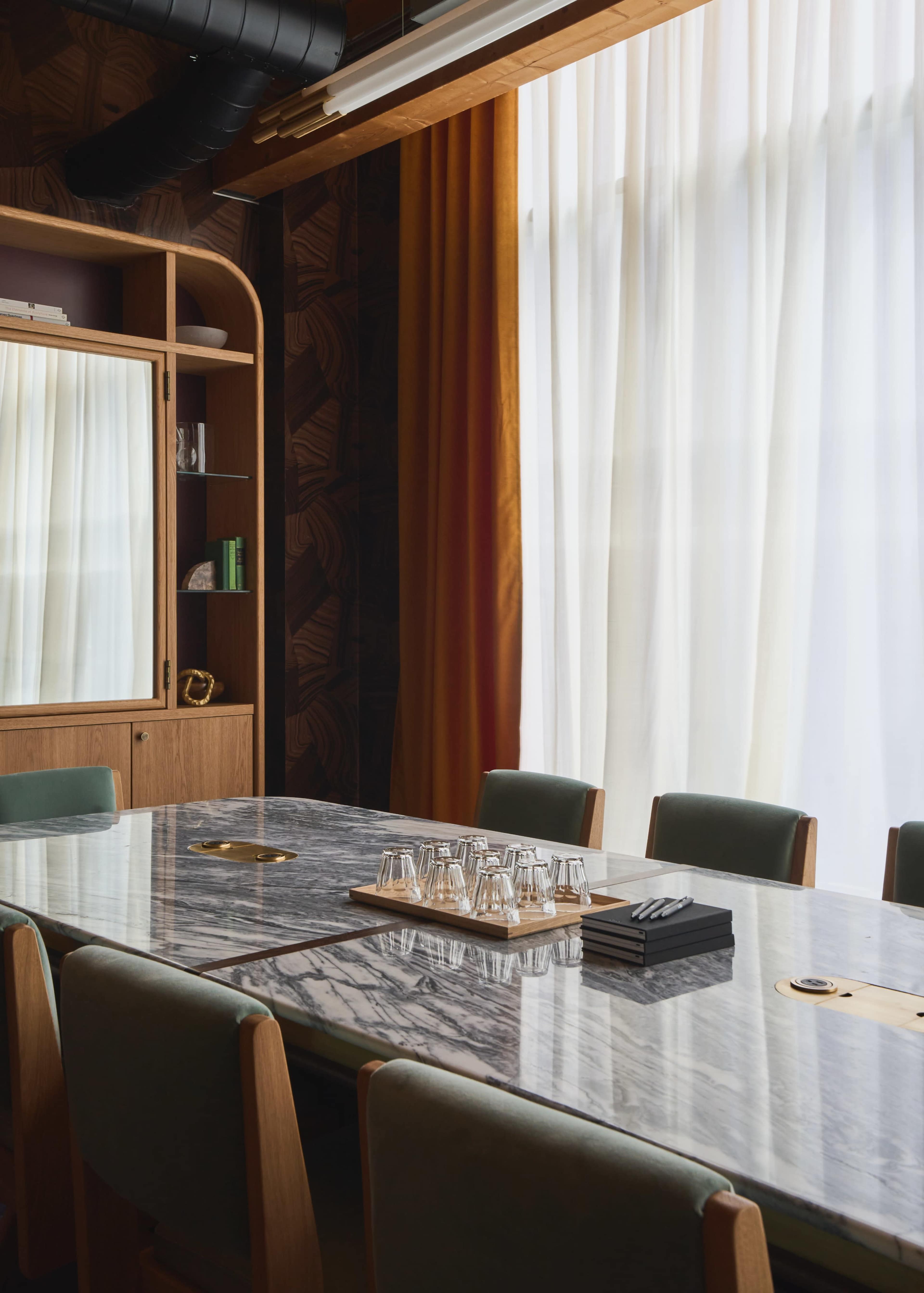 A modern conference room featuring a large marble table surrounded by green upholstered chairs, with floor-to-ceiling windows draped in sheer white curtains.
