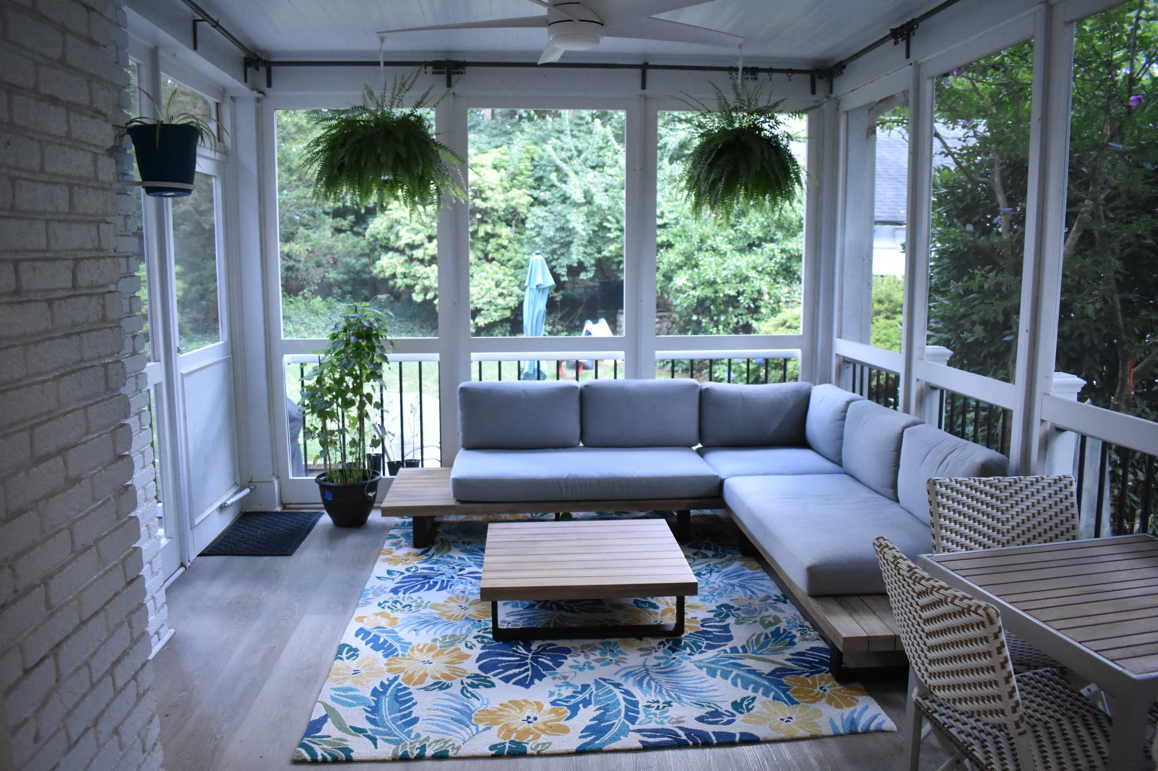 The image shows a screened porch with a gray sectional sofa, a wooden coffee table, potted plants, and a floral rug, with a view of a green landscape outside.