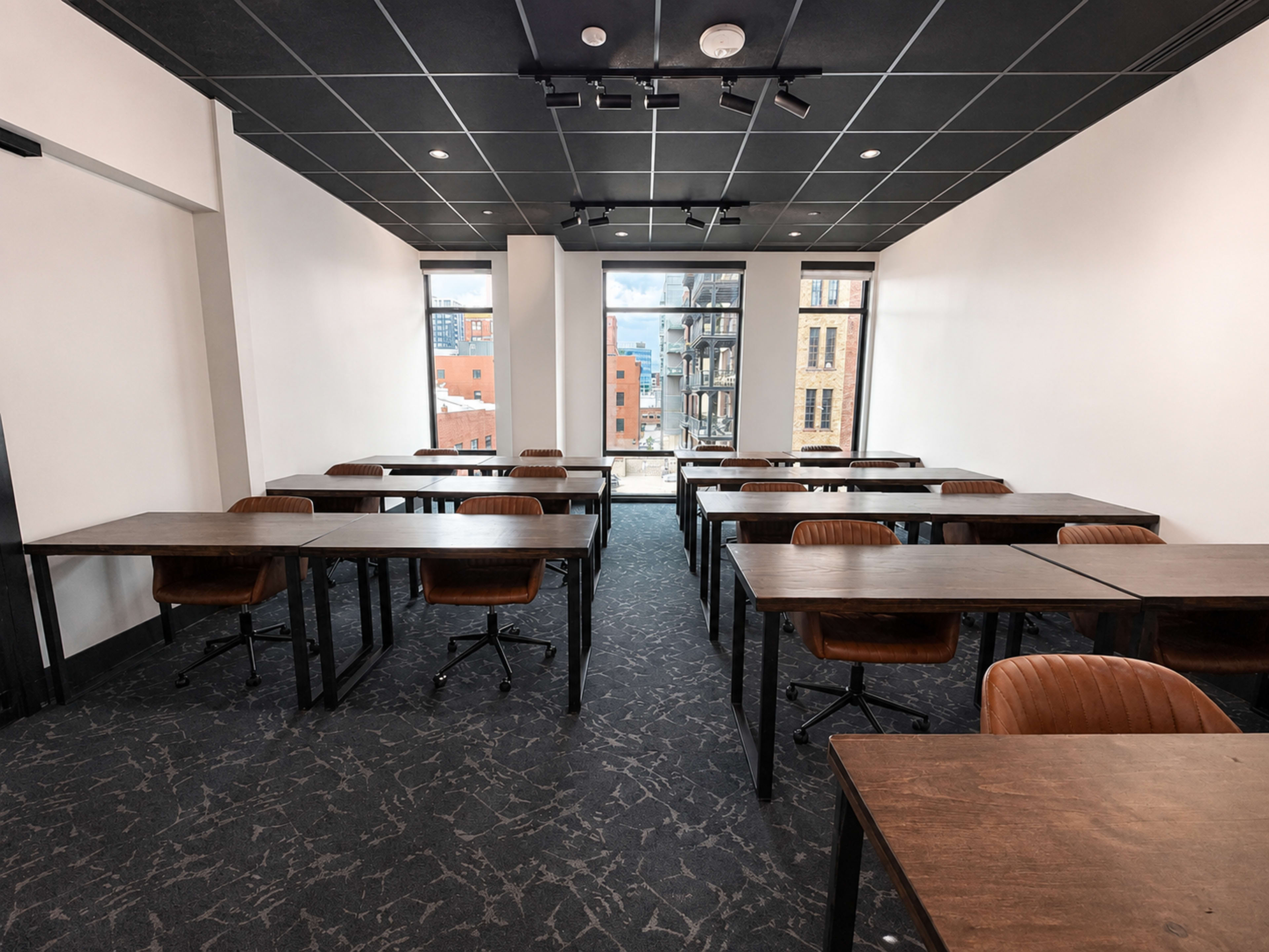 The image shows a modern classroom with several rows of wooden tables and chairs arranged neatly, and large windows providing a view of outside buildings.