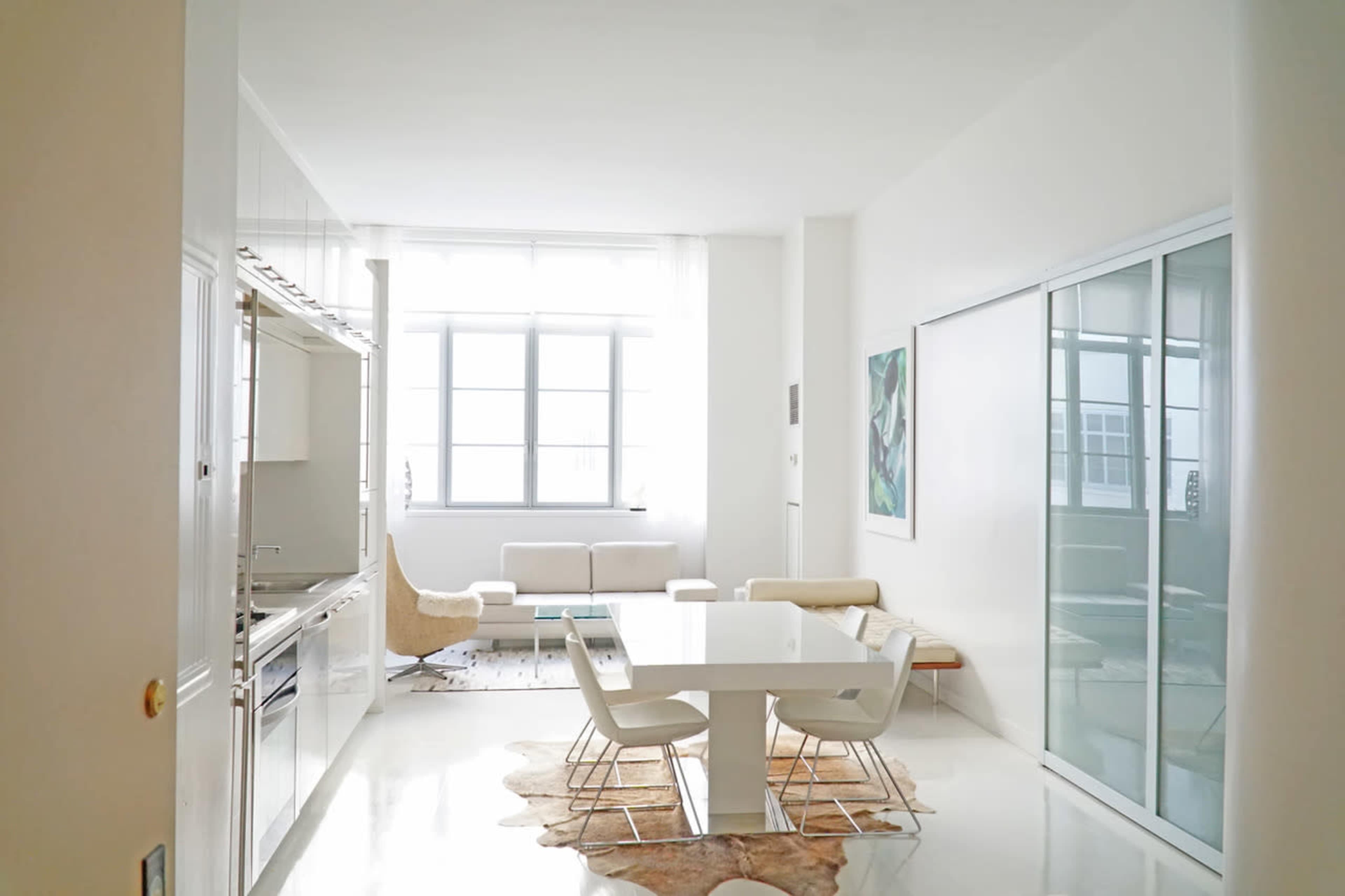 A modern kitchen and dining area with a glass wall, a white dining table, and a light-filled living space featuring a sofa and decorative artwork.