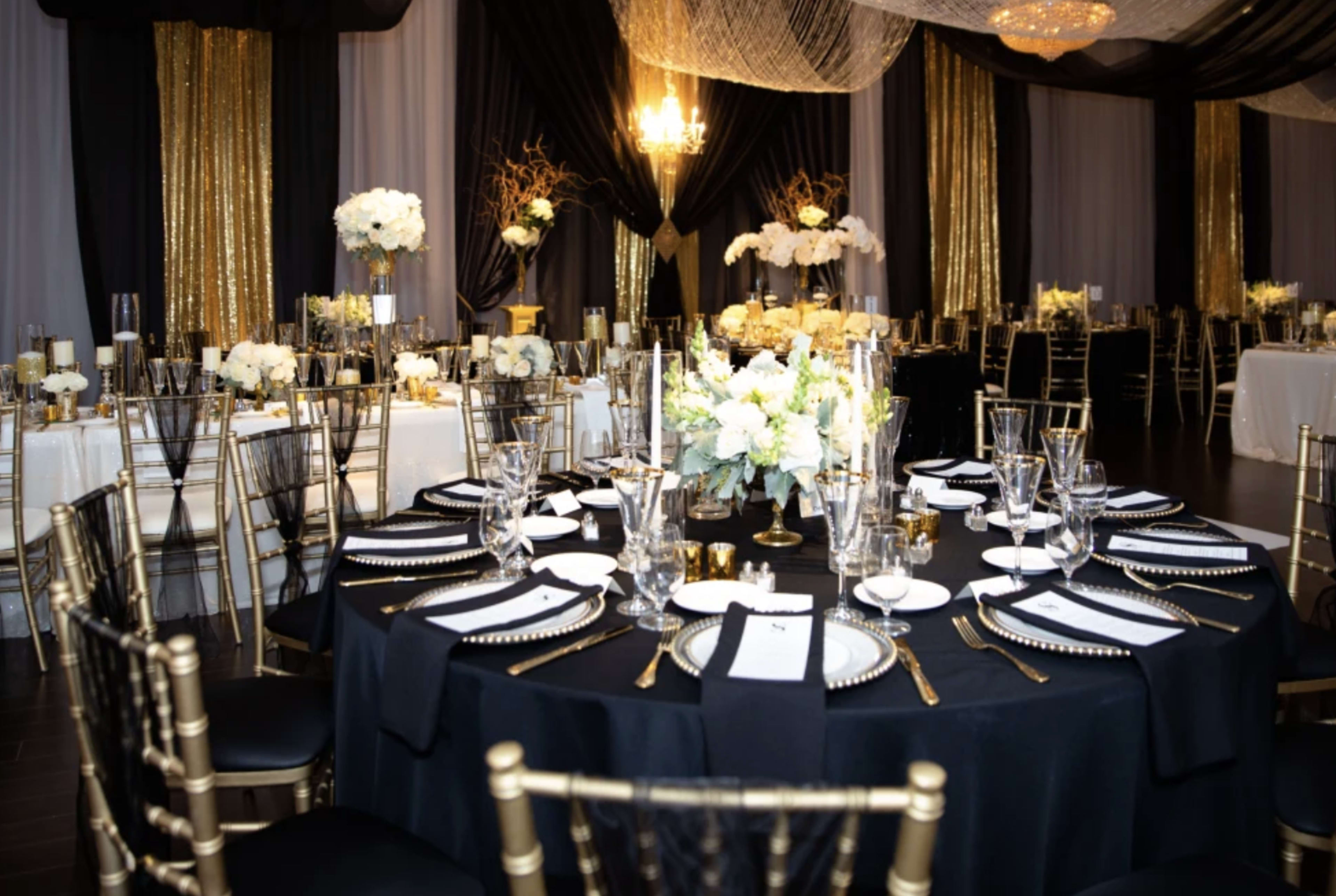 A banquet hall features elegantly arranged tables with black and gold decor, white floral centerpieces, and shimmering drapes.