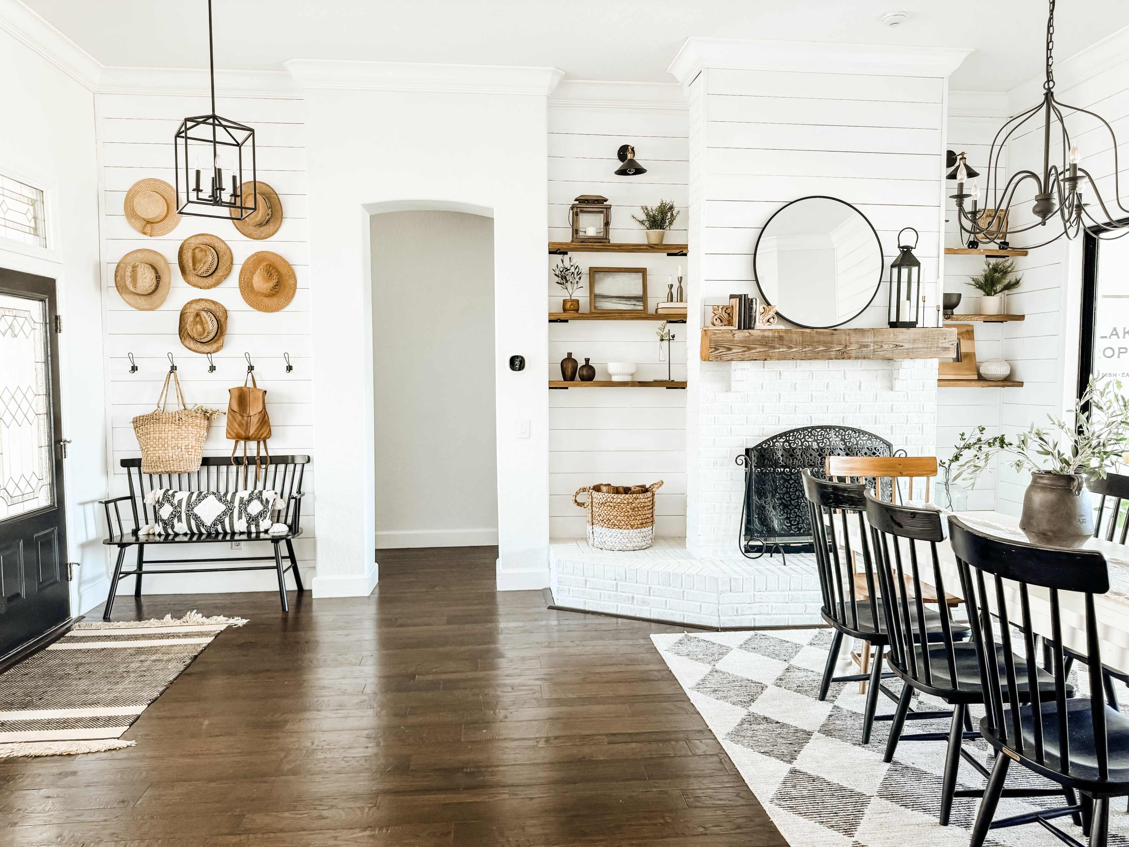 The image shows a spacious, well-lit room featuring a dining area with a patterned rug, a black table surrounded by chairs, and a cozy fireplace with decor and seating elements nearby.