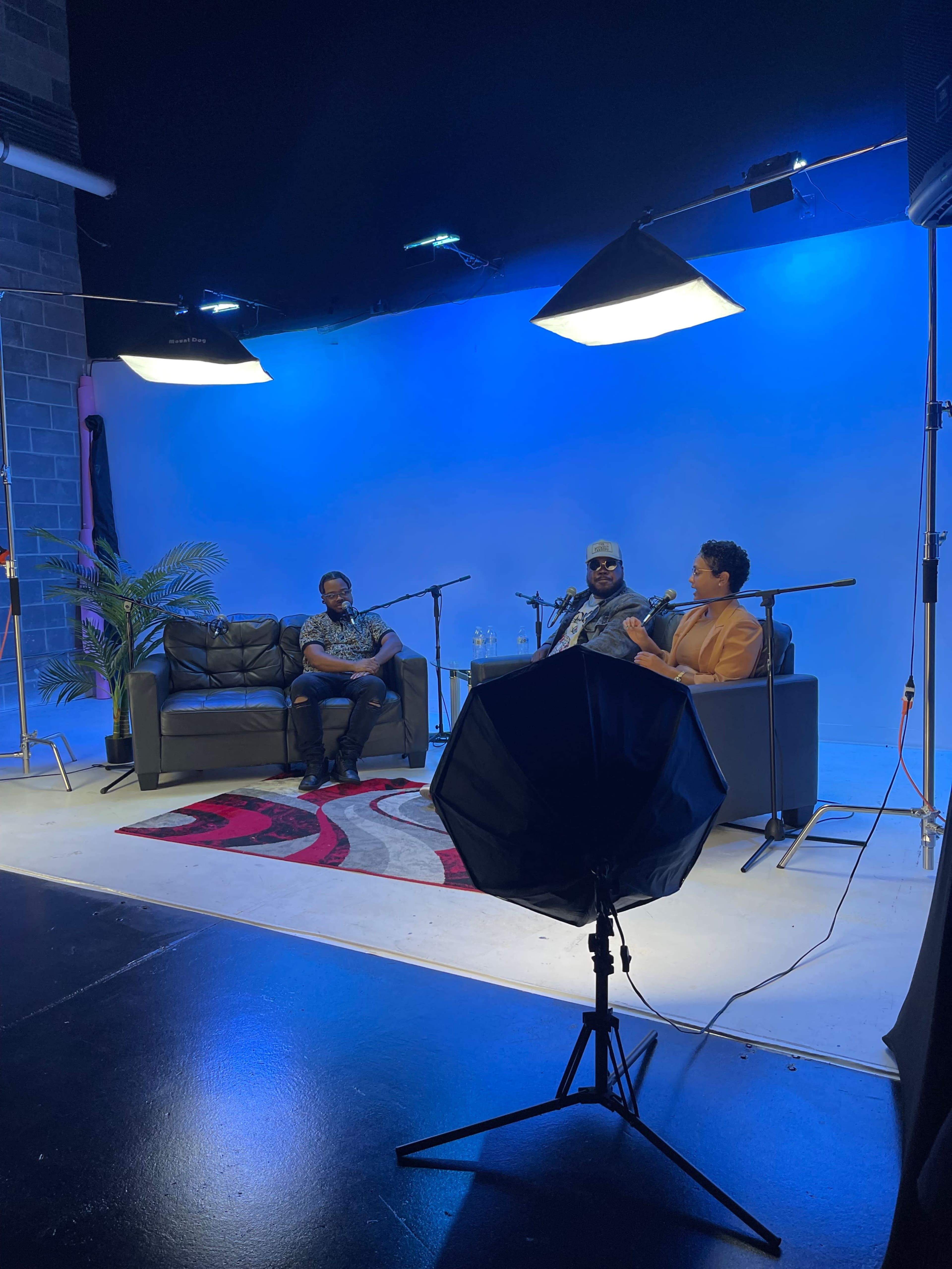 A three-person discussion is taking place on a set with blue lighting, featuring two speakers seated on a couch and a third person in the background, with microphones positioned for recording.