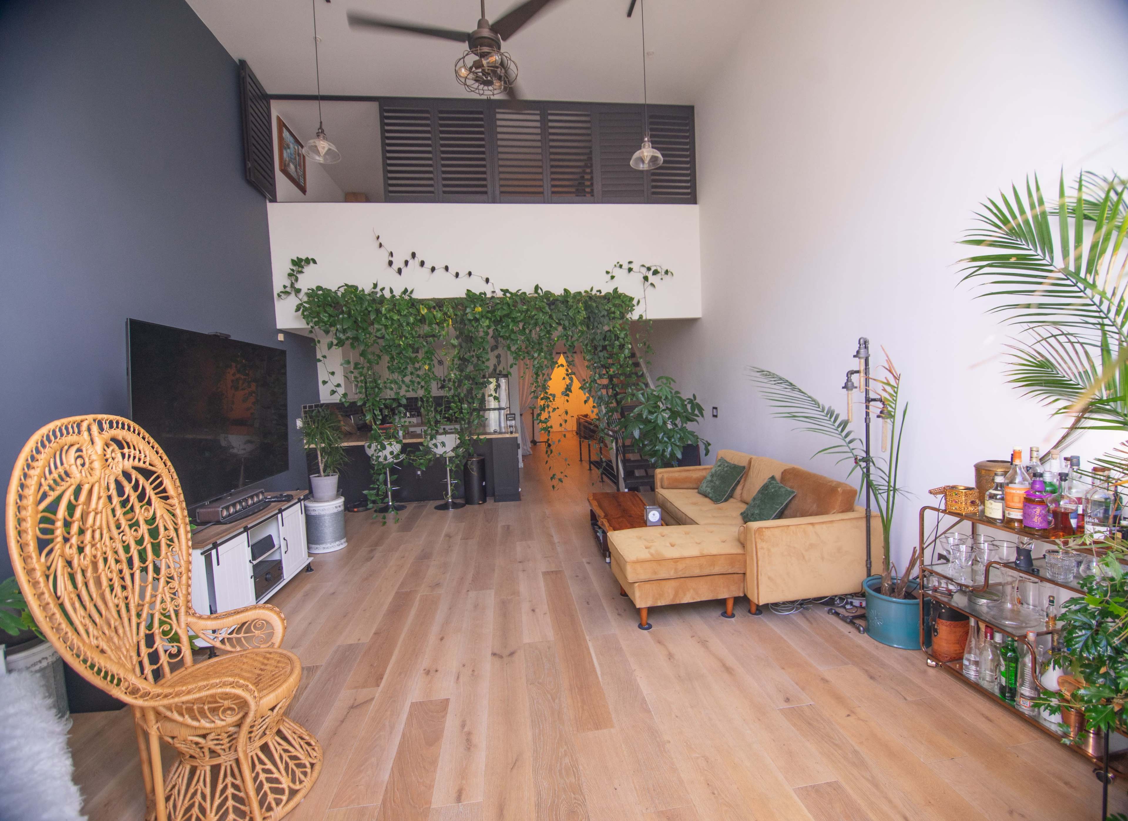 The room features a spacious layout with wooden flooring, a large television, a yellow couch, and greenery hanging from the walls.