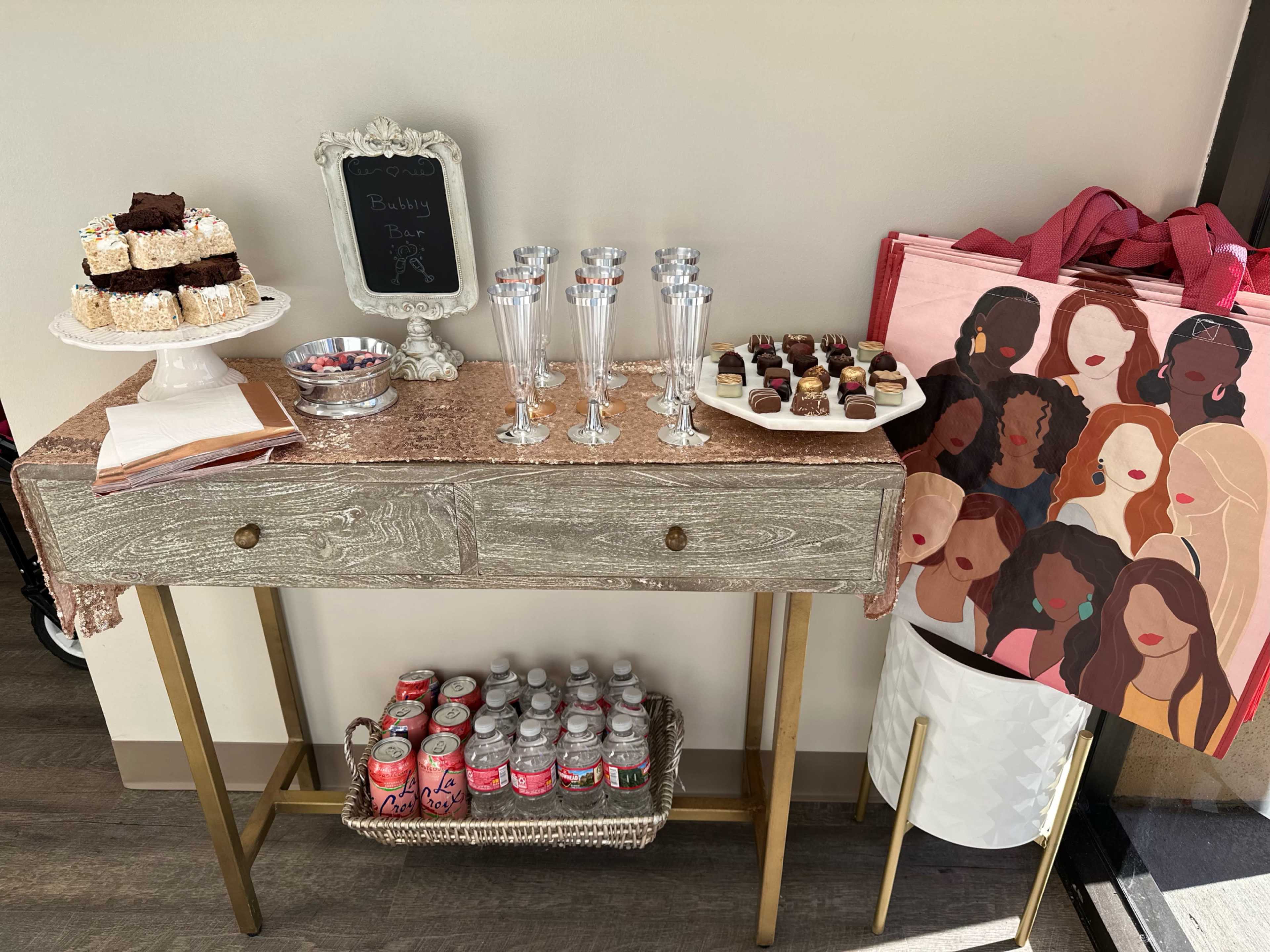 A table decorated with a pink cloth features desserts, sparkling glasses, and a decorative bag, set against a wall.