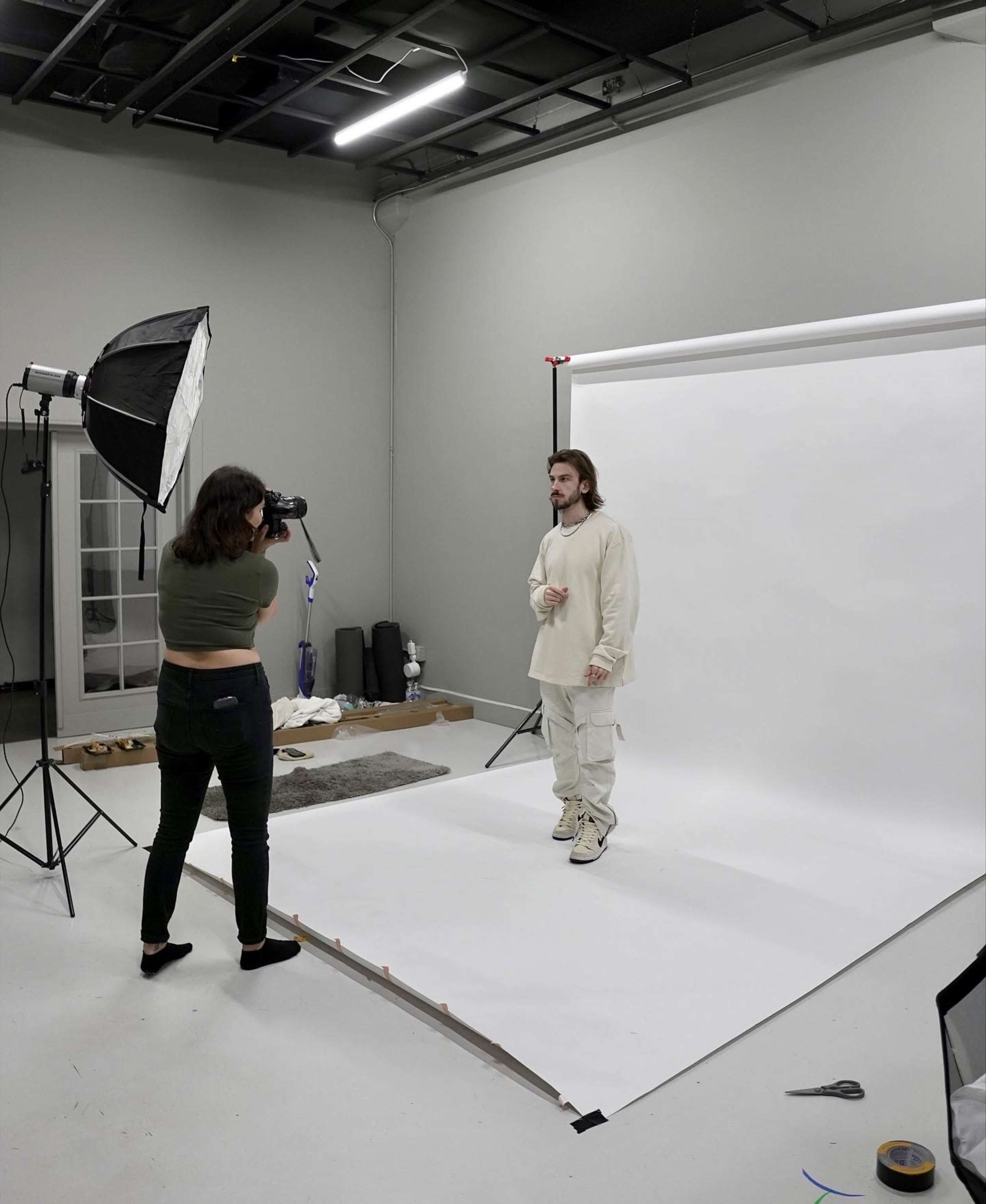 A photographer shoots a subject wearing light clothing against a plain white backdrop in a studio setting.