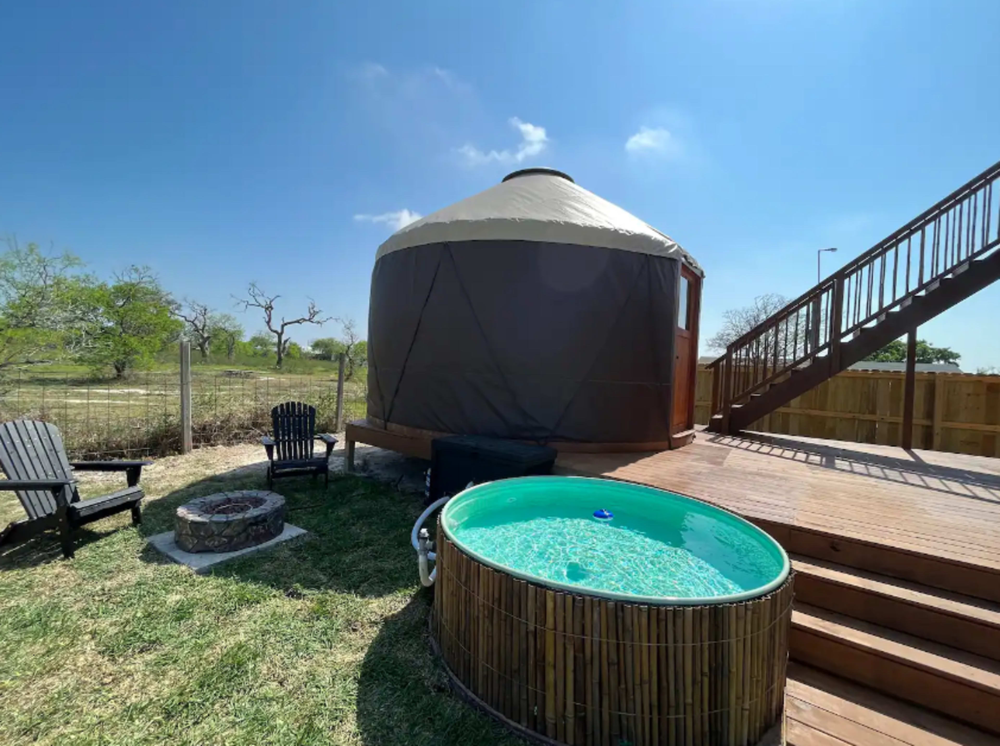 A yurt is situated beside a small above-ground hot tub on a deck overlooking a grassy area.