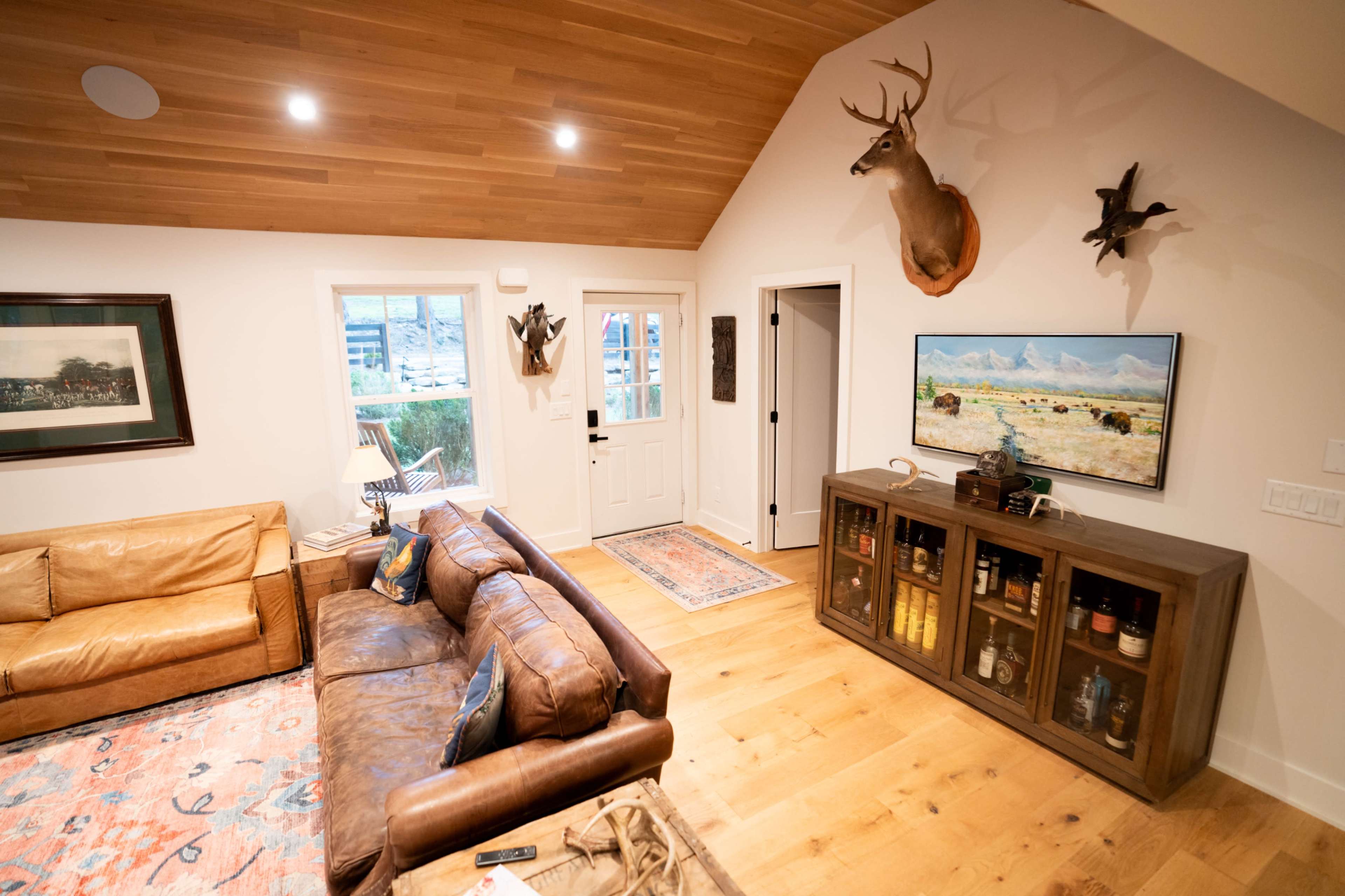 The cozy living room features a mix of leather seating, a mounted deer head, and a cabinet displaying various items, with a view of an entrance door.