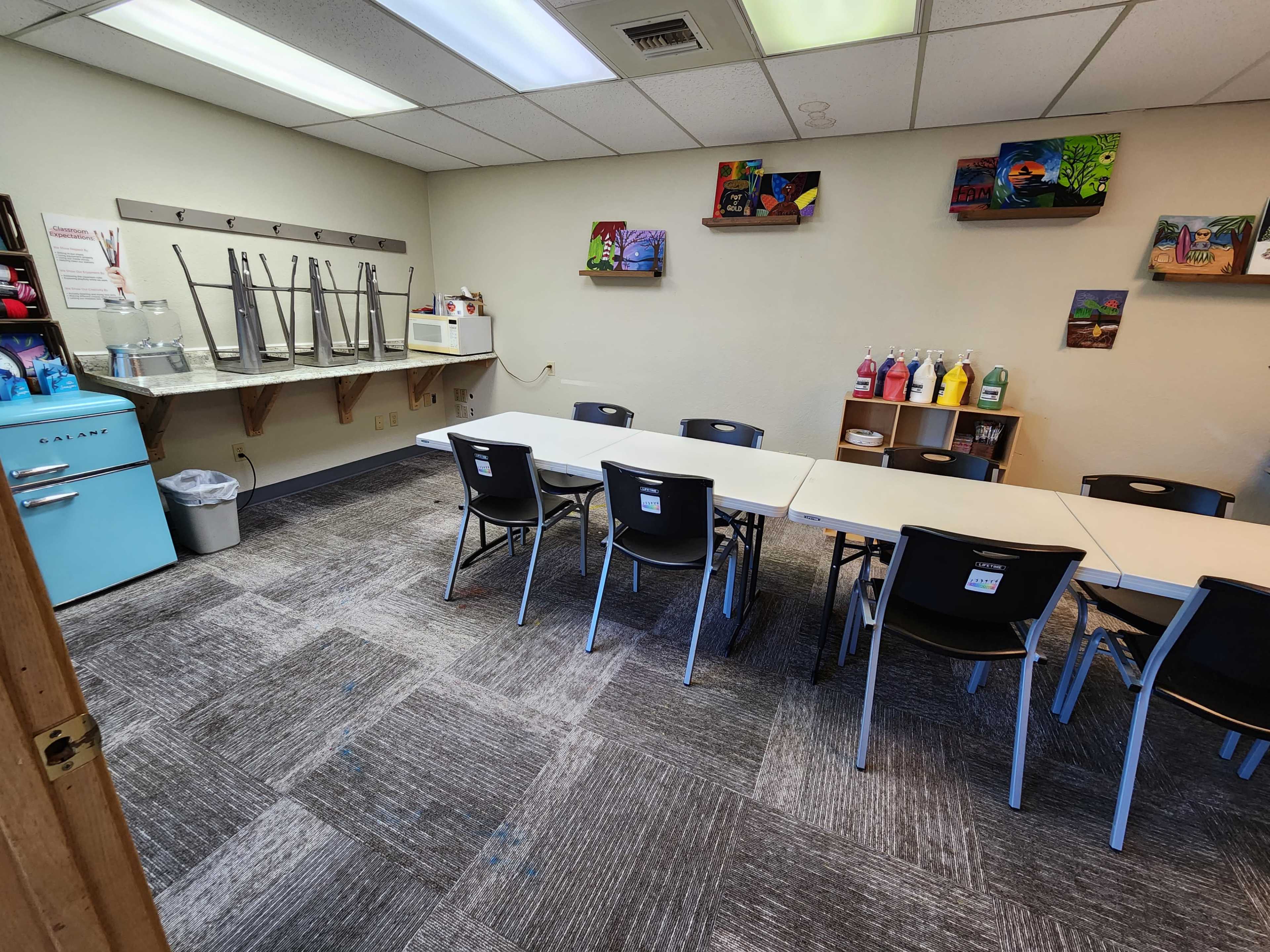 A classroom with a long table and black chairs arranged in rows, a blue retro refrigerator, and shelves displaying colorful bottles and artwork on the walls.