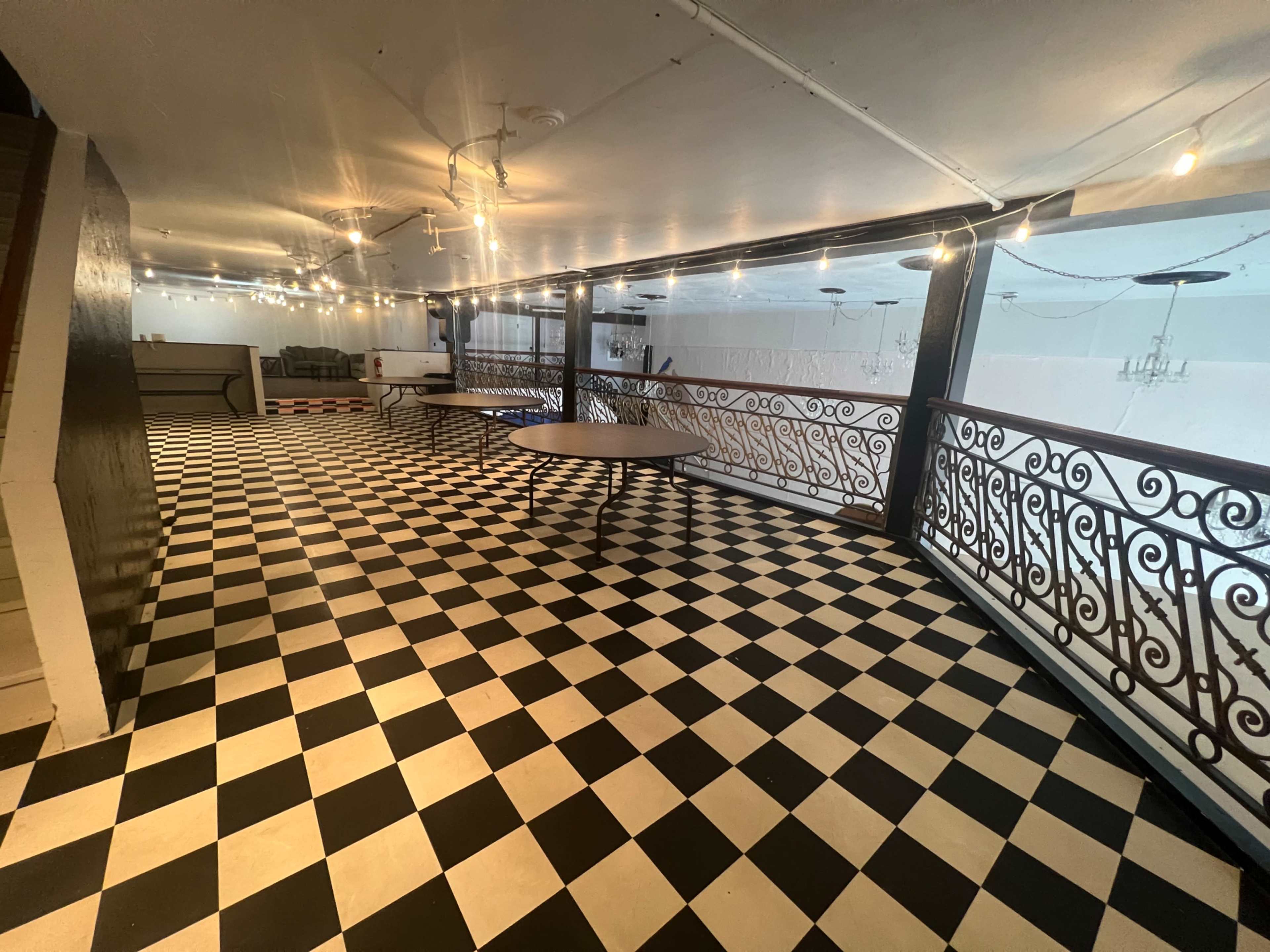 The image shows a spacious balcony area with a black and white checkered floor, decorative iron railings, and overhead string lights.