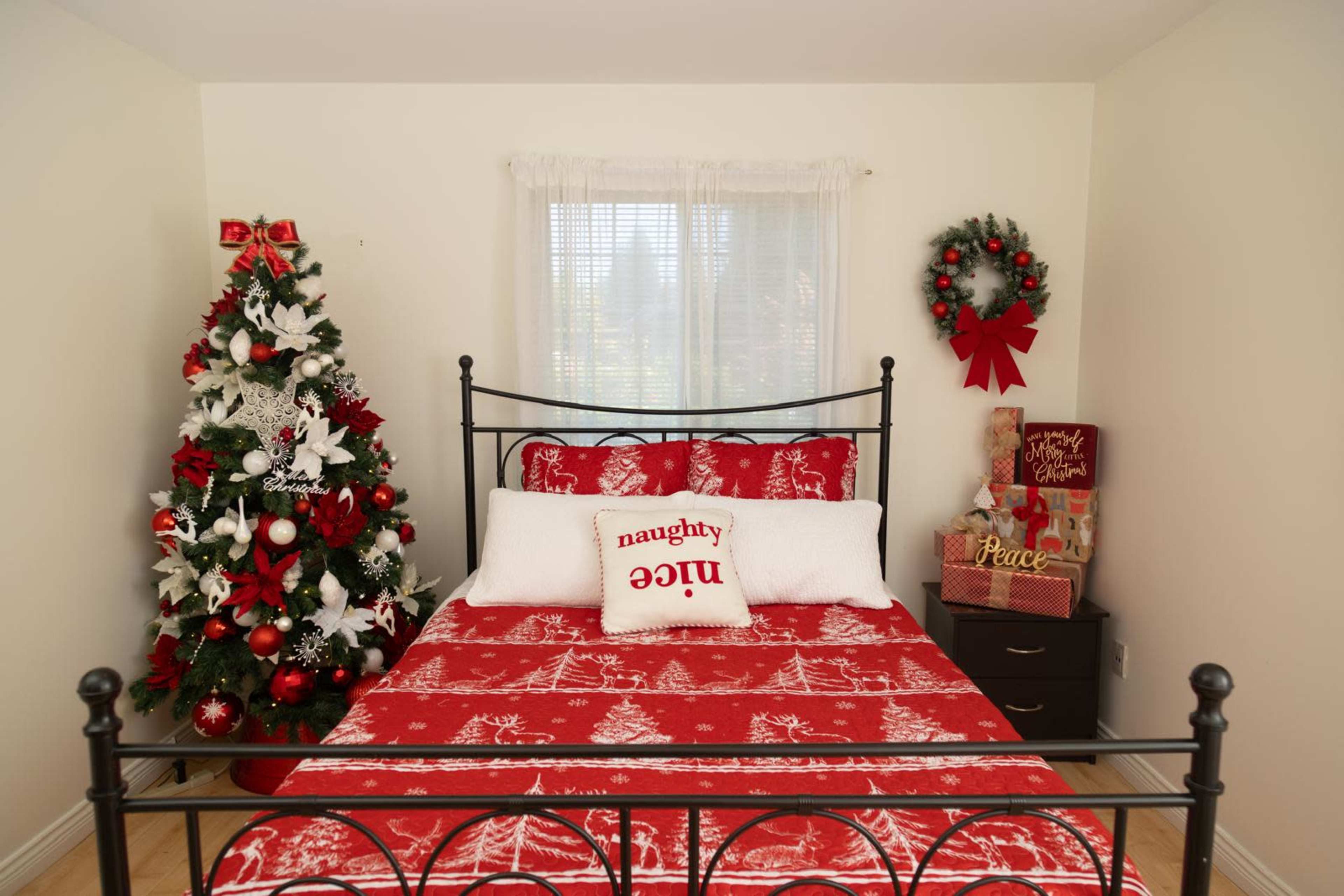 The image features a decorated bedroom with a red-themed bedspread, featuring a sign that reads "naughty," accompanied by a Christmas tree adorned with white and red decorations.