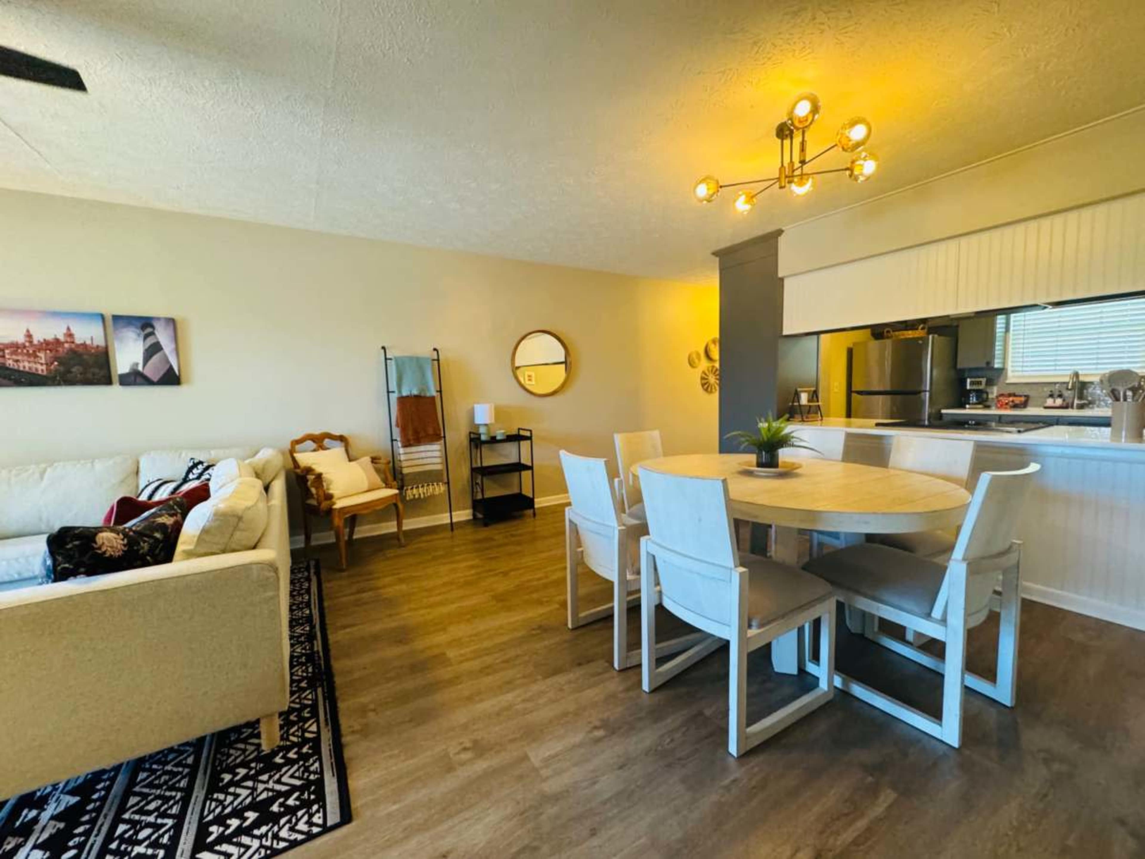 The image shows a modern living and dining area featuring a beige couch, a round dining table with white chairs, and kitchen appliances visible in the background.