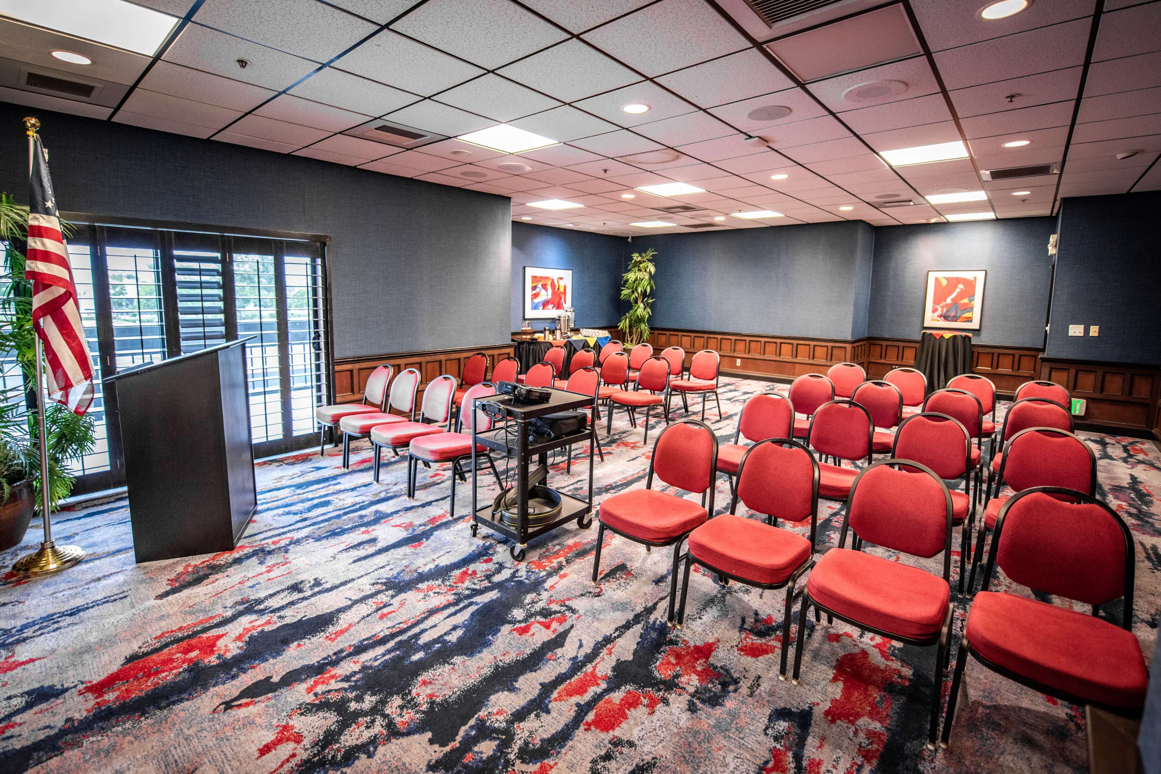The room features rows of red chairs arranged for a presentation, with a podium, a projector on a stand, and an American flag in the corner.