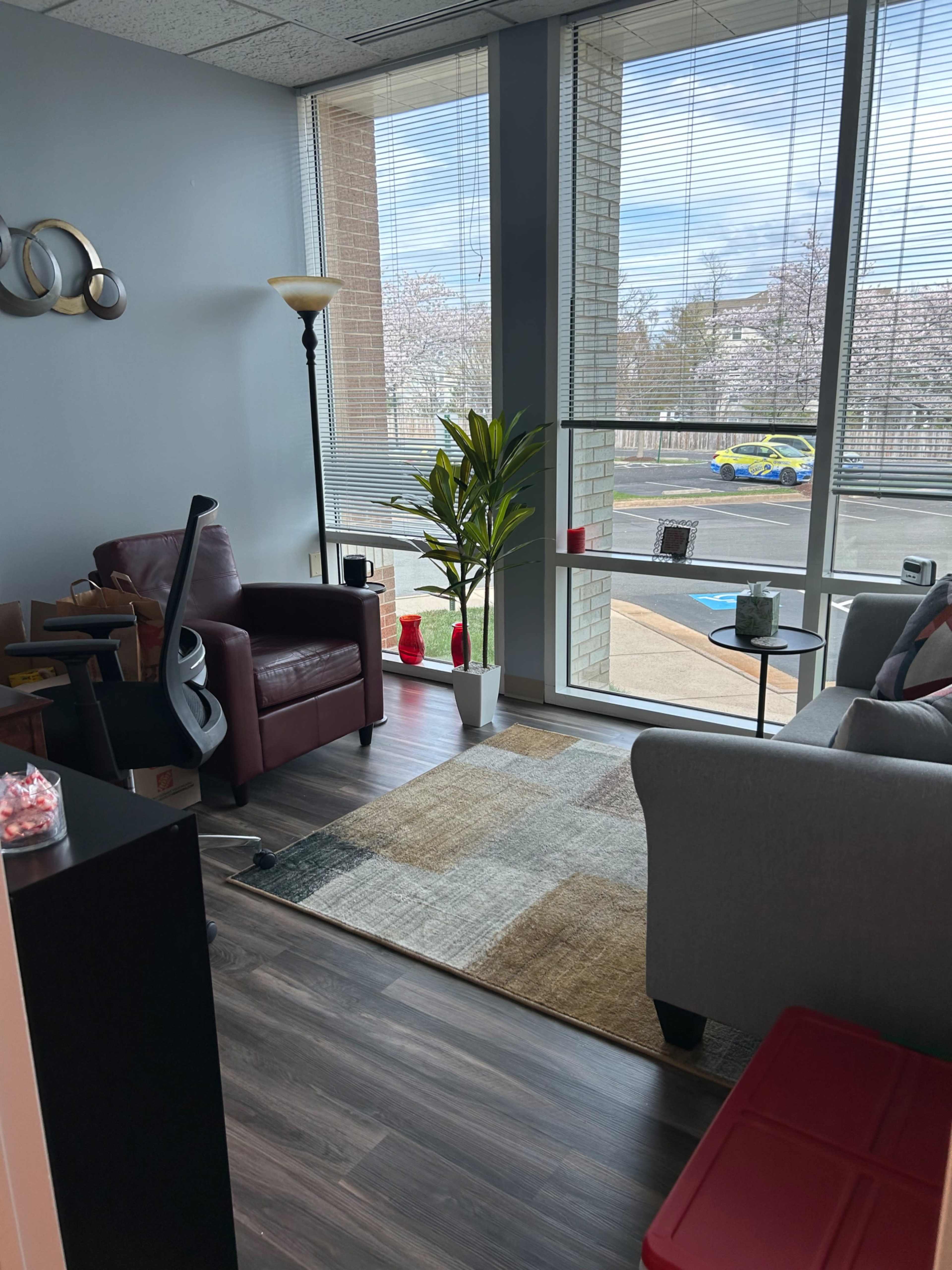 The image shows a modern office space featuring a plant, seating areas with a red chair and gray couch, and large windows letting in natural light.