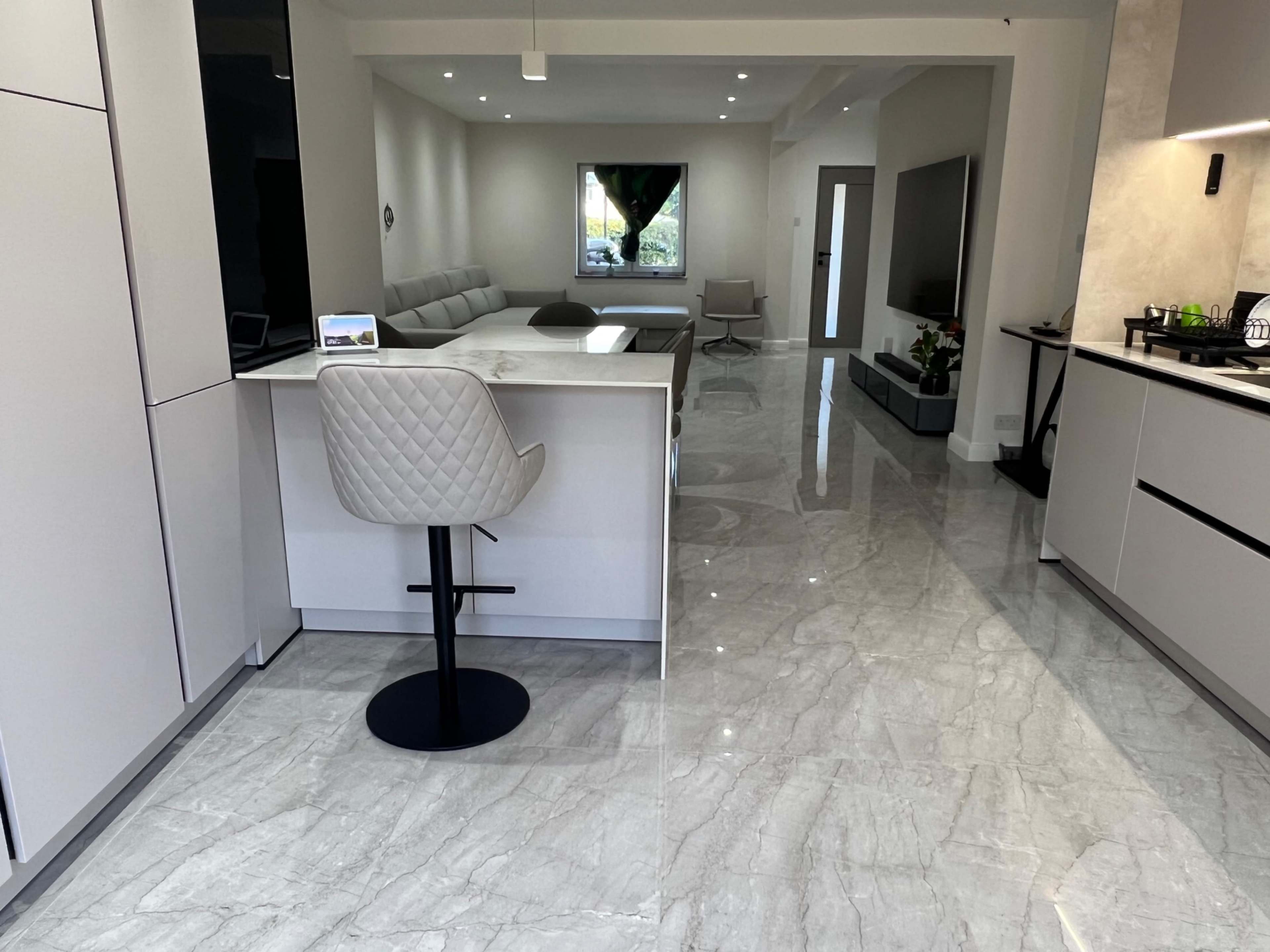The image shows a modern kitchen and living area with a marble floor, an island with a stool, and a seating area visible in the background.