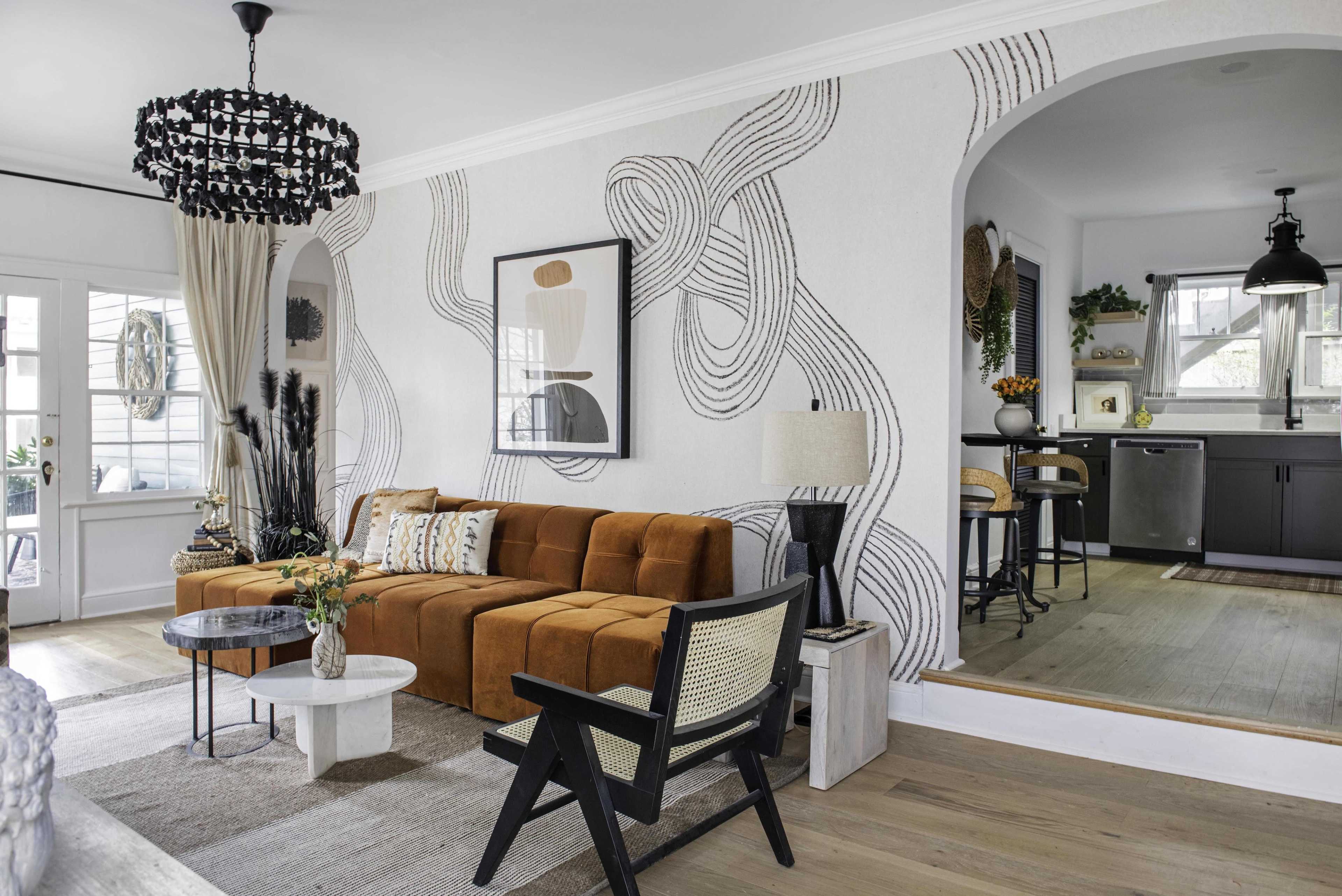 The image shows a modern living room featuring a brown couch, a white coffee table, and a decorative wall with abstract line art, adjacent to a kitchen area.
