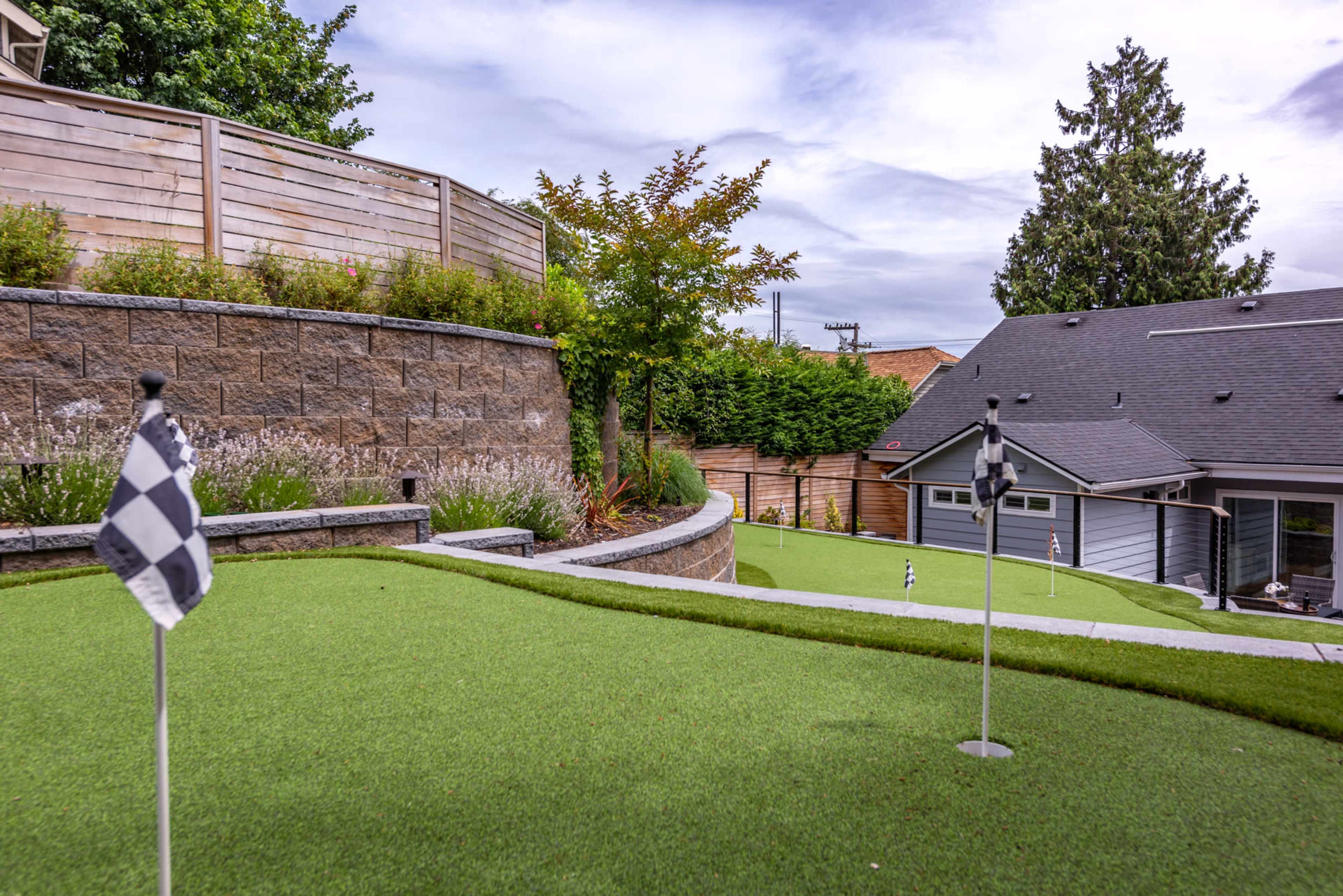 A landscaped backyard with artificial turf, featuring two small golf holes marked by flags and a hillside garden in the background.