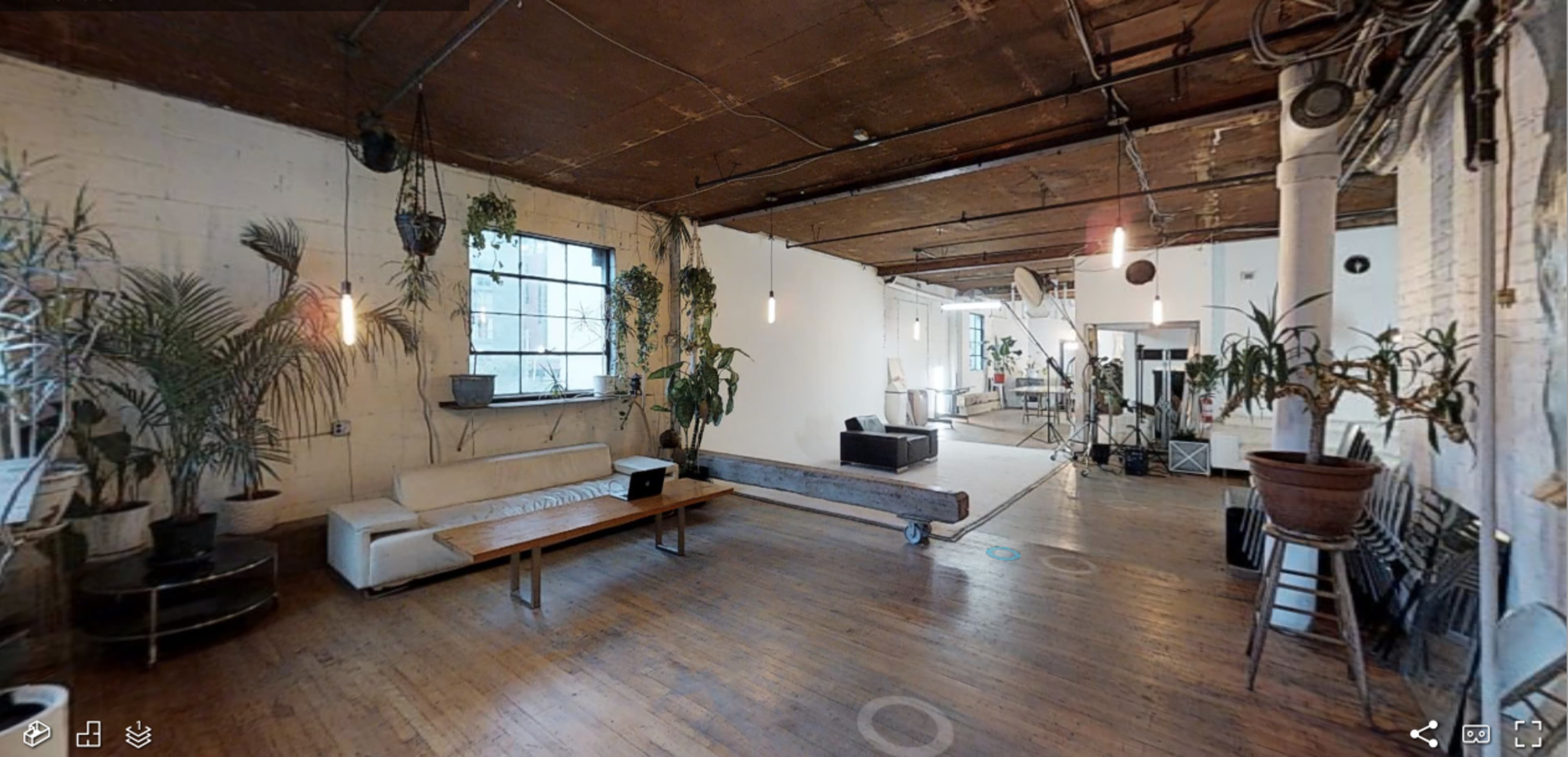 The image shows an industrial-style loft space with wooden floors, a large window, potted plants, a sofa, and a minimalist decor featuring a staircase in the background.
